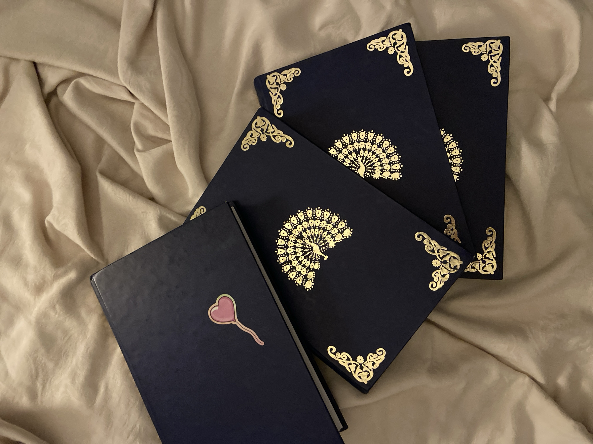 Four dark blue hardback books are displayed on a crumpled light beige fabric surface. The uppermost book, positioned horizontally on the left, bears a pink heart-shaped lollipop sticker on its cover. Below it, three additional dark blue books are fanned out, overlapping towards the lower right. Each visible cover of these lower books features a gold foil design: a central peacock-feather-like emblem and ornate corner scrollwork. No individuals or actions are depicted. The environment consists solely of the fabric texture and the books. The scene is situated in Budva, Montenegro.