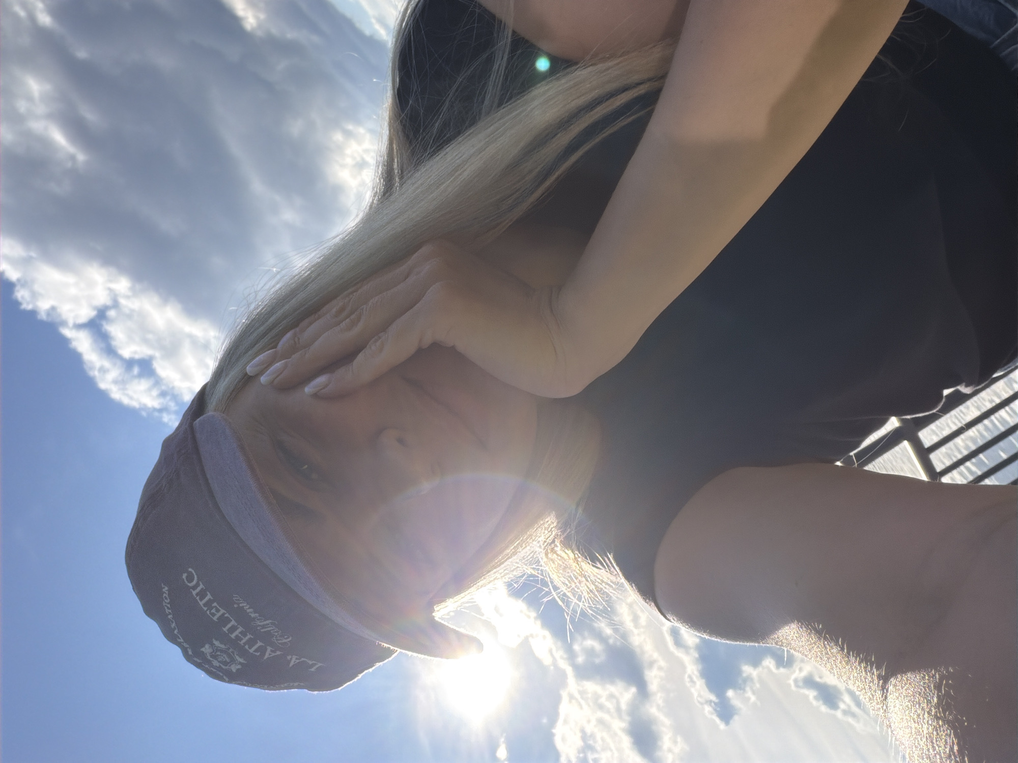 A woman with blond hair is holding her hand to her face, obscuring her eye. She is wearing a dark shirt and a baseball cap. The cap is light brown with the inscription "LA ATHLETIC California" on it. The background consists of a blue sky with white clouds. The sun is shining brightly, creating a lens flare. The location cannot be determined from the image.