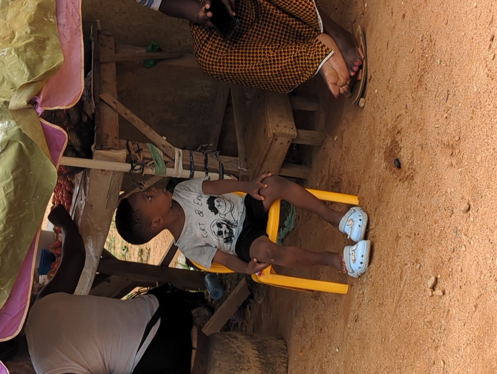 The image shows a young boy sitting on a yellow plastic chair in what appears to be a small, makeshift structure. The boy is wearing a light-colored t-shirt with a graphic print, black shorts, and light blue shoes. He is looking to his left. A person is seated behind him, partially obscured, wearing a white t-shirt and black pants. Another person, wearing a patterned yellow and brown dress, is seated above the boy, holding a mobile phone. The scene is likely in Sabon Tasha, Nigeria, based on the context, and the ground is covered in reddish-brown dirt. A rudimentary wooden structure surrounds the child and adults, and a tarp is visible above.