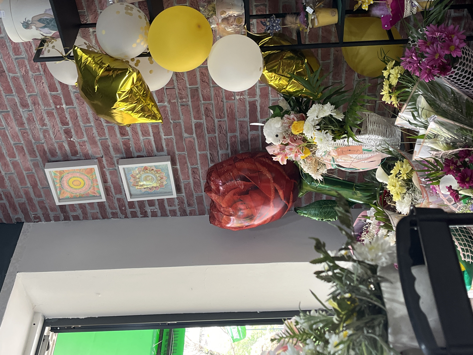 The image shows an interior scene with decorations suggesting a celebratory event or flower shop setting. The backdrop features a brick wall, against which various balloons (white, yellow, gold, and red) are arranged. There are also framed, possibly art-related, objects on the wall. To the right of the image, bouquets of flowers are prominent. The bottom of the image shows a window with a view of greenery outside. The location is potentially Burgas, Bulgaria, but there isn't explicit evidence of this in the image.