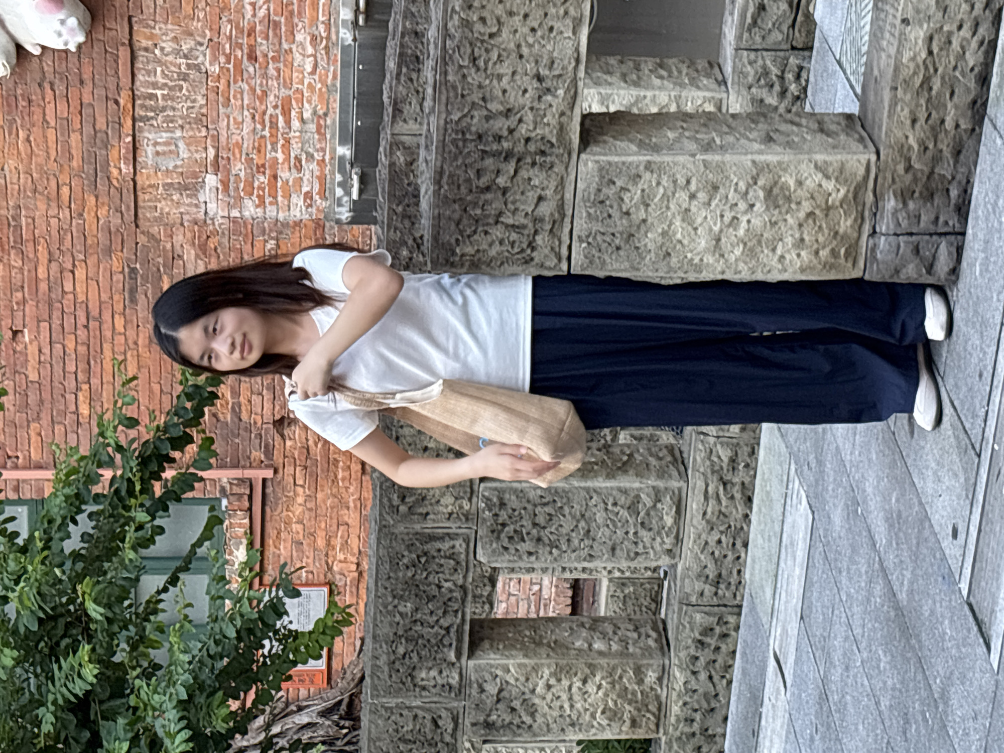 A young woman with long, dark hair stands on a paved walkway in front of a textured brick wall. She is wearing a white t-shirt and dark, wide-leg pants. A light-colored woven tote bag is slung over her right shoulder. She is looking to her left, with her head tilted slightly. Behind her, the brick wall has a green-trimmed window and a small, orange plaque with white text. A tree with green foliage is visible on the left side of the image. In front of the wall, there is a stone structure with rough-hewn blocks and openings. The ground is paved with gray square tiles. A portion of a white, plush figure with pink paws is visible at the top right of the image, attached to the brick wall. The scene appears to be outdoors, possibly in an urban area. This scene is located in the Yuzhong District of China.
