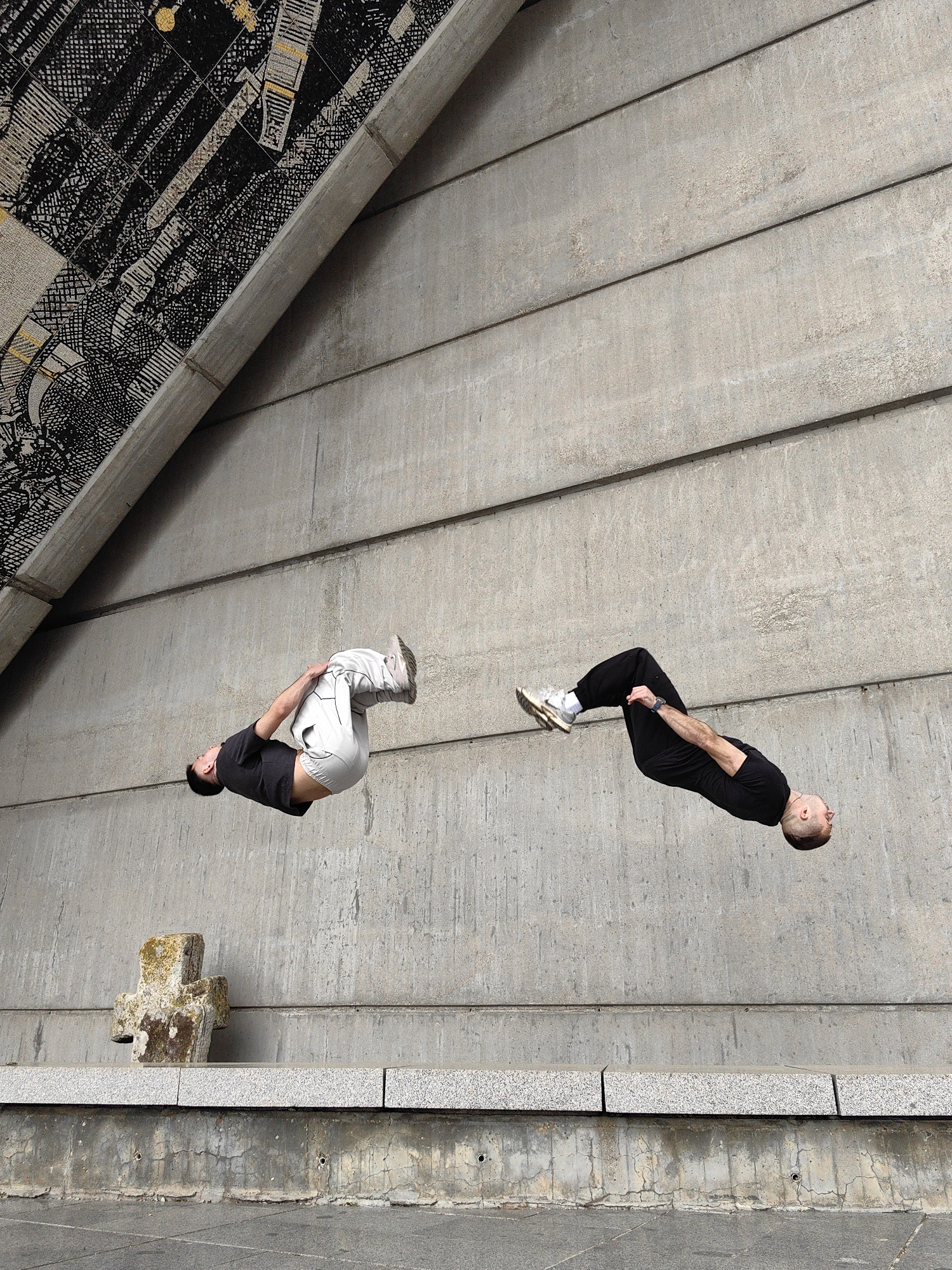 The image captures a dynamic scene featuring two men performing aerial maneuvers against a stark, architectural backdrop. Both individuals are suspended mid-air, appearing to execute flips or somersaults. The man on the left, wearing a dark t-shirt and light-colored pants, is in a tucked position with knees drawn towards his chest. The man on the right, dressed in black, is in a more extended, slightly arched posture. Their synchronized yet distinct movements suggest high-level athletic activity like parkour or freerunning.
The setting is dominated by large concrete structures. The background consists primarily of a tall, light gray concrete wall, constructed from distinct horizontal panels, showing some weathering and minor staining. Above this wall, an angled overhead structure features an intricate mosaic art piece. This mosaic is predominantly black and white, with complex, detailed patterns, and includes subtle accents of gold or yellow, possibly depicting abstract or symbolic imagery.
Below the aerial figures, a robust stone ledge or platform is visible. Resting on this ledge, towards the left, is a weathered stone cross, partially covered with moss, suggesting age and a commemorative purpose. Further below the ledge, there is a darker concrete base with visible cracks and small holes, leading down to a tiled ground surface. The lighting indicates a bright, clear daytime, with even illumination and no strong shadows. No text is visible in the image. The composition effectively contrasts the fluid human forms with the rigid, geometric lines of the brutalist-style architecture and the detailed texture of the mosaic.