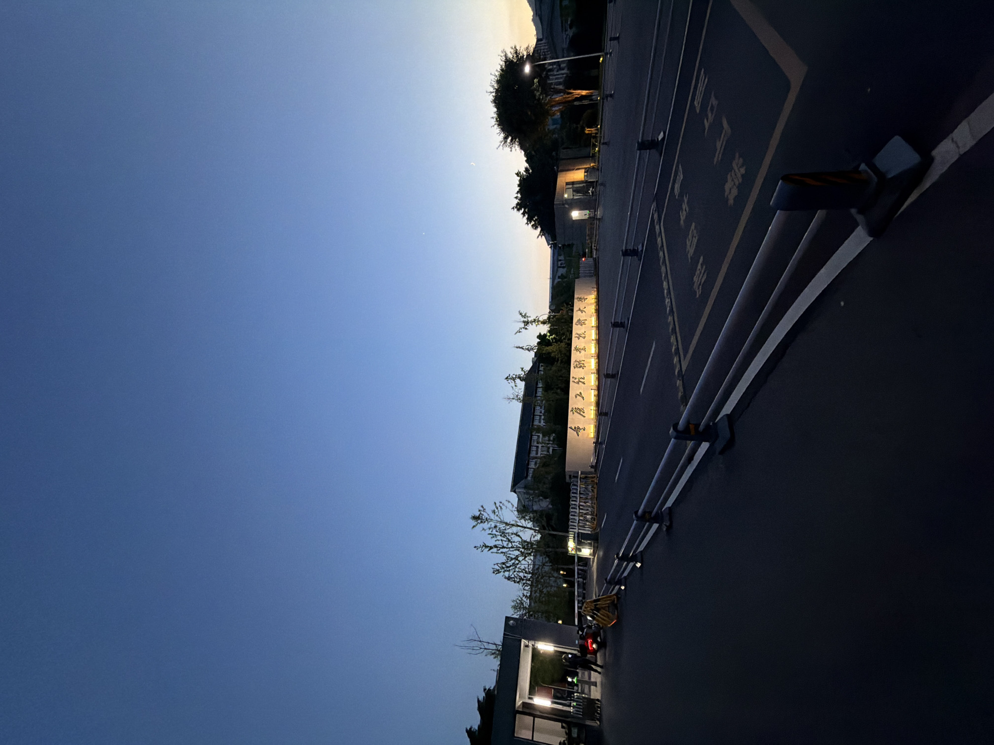 This image depicts the entrance of Chongqing University of Technology at Jiangjin Campus, China, during dusk. The sky is a deep blue, transitioning to a lighter orange hue at the horizon on the right side, with a visible crescent moon and a small star. In the foreground, a dark asphalt road stretches towards the entrance, marked with yellow lines and symbols, including "禁止停车" (No Parking). Metal barriers with black and yellow reflective posts line the sides of the road.
To the left, a modern entrance gate structure with overhead lighting is visible. A person is standing near a scooter at the gate. Further back, a curved illuminated wall displays Chinese characters, which read "重庆理工大学江津校区" (Chongqing University of Technology Jiangjin Campus). Beyond the entrance, buildings of the university are visible, characterized by their darker facades and illuminated windows. Trees and streetlights are scattered throughout the campus grounds. The overall scene suggests a quiet, early evening at the university.