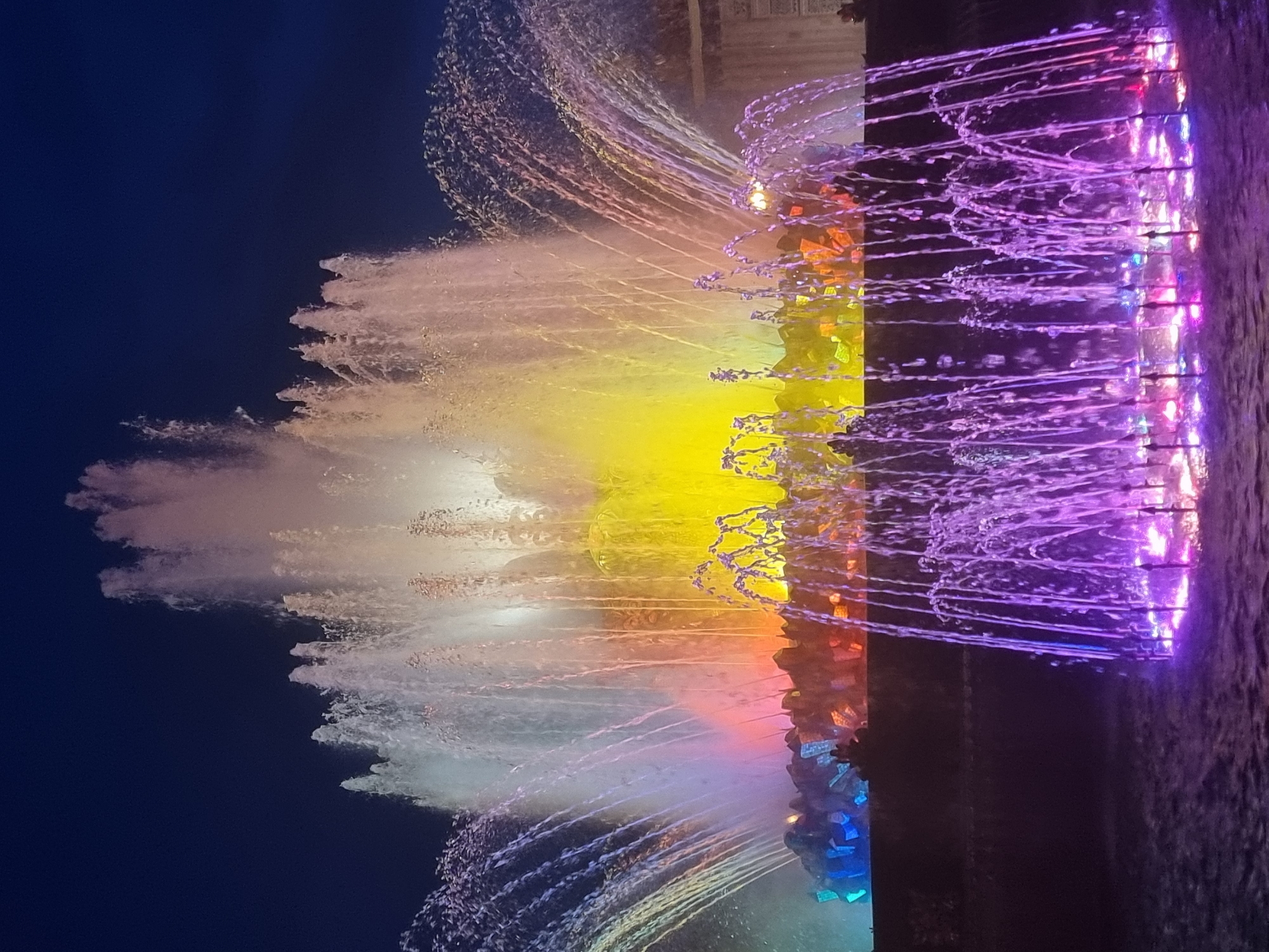 A night scene depicts a large, multi-tiered fountain in Moscow, Russia, illuminated by vibrant colored lights. The water jets spray upwards and outwards, catching the light and creating a dynamic display. The foreground features a lower section of the fountain with numerous fine water streams illuminated in purple and pink, originating from the water's surface. Behind this, a larger section of the fountain rises, with thicker plumes of water lit by yellow and orange lights, and a softer white light at the very top. The base of this central section appears to be adorned with rocks or sculpted elements, also lit with various colors, including blue, purple, and orange. The background is a dark, clear night sky. The overall impression is one of a spectacular, illuminated water feature in a park setting.