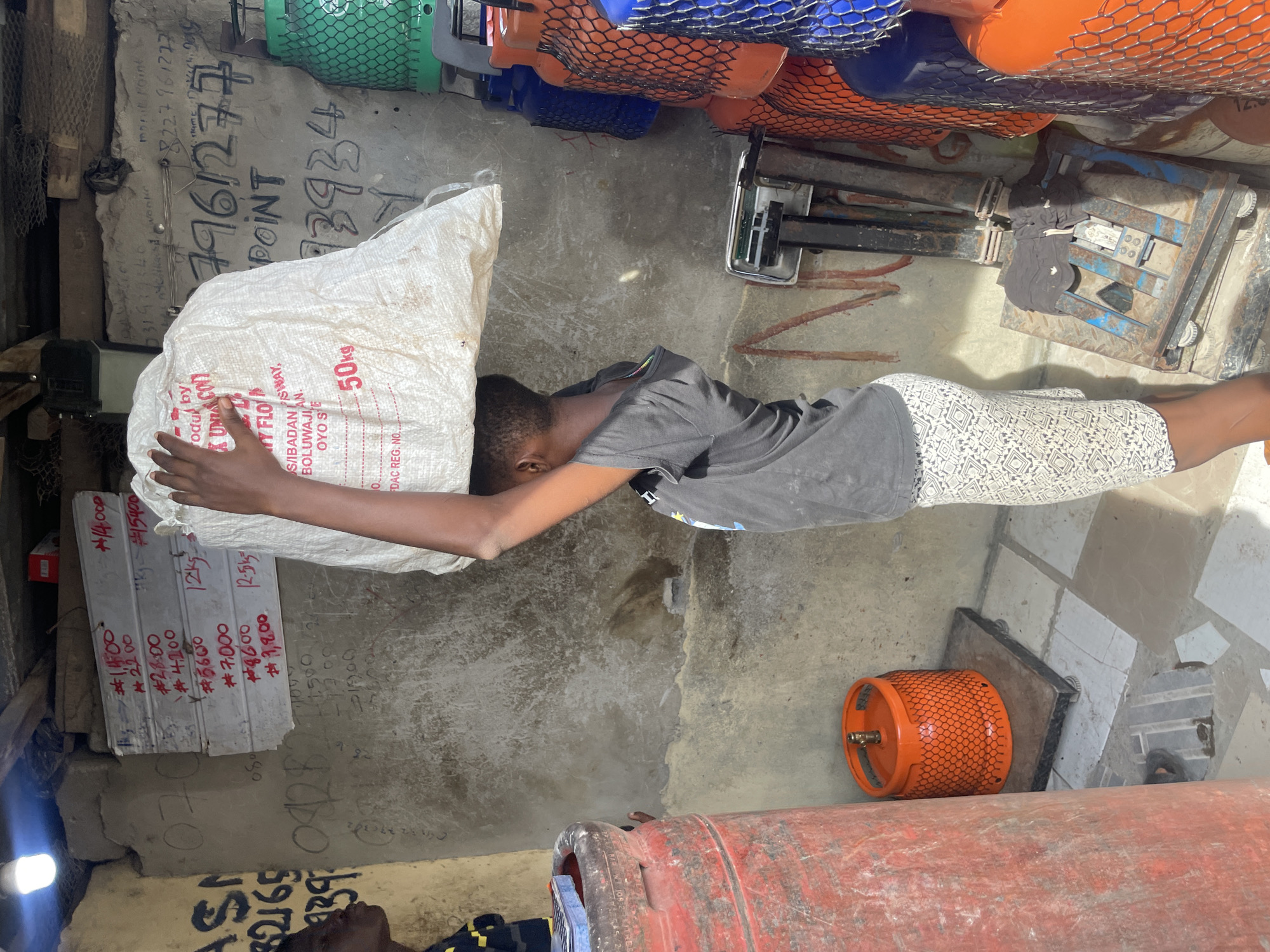 Here is a description of the image:
The image shows a young person lifting a large, white sack, labeled "50kg," in what appears to be a small business or storage area in Ado, Nigeria. The person is wearing a grey t-shirt and patterned shorts. Various items surround the person, including stacks of price-marked boards on the wall to the left, gas cylinders in orange and other colors, and a weighing scale. Text is visible on the walls, including phone numbers and business-related information. The sack has the word "FLOUR" and business information indicating "S/IBADAN."