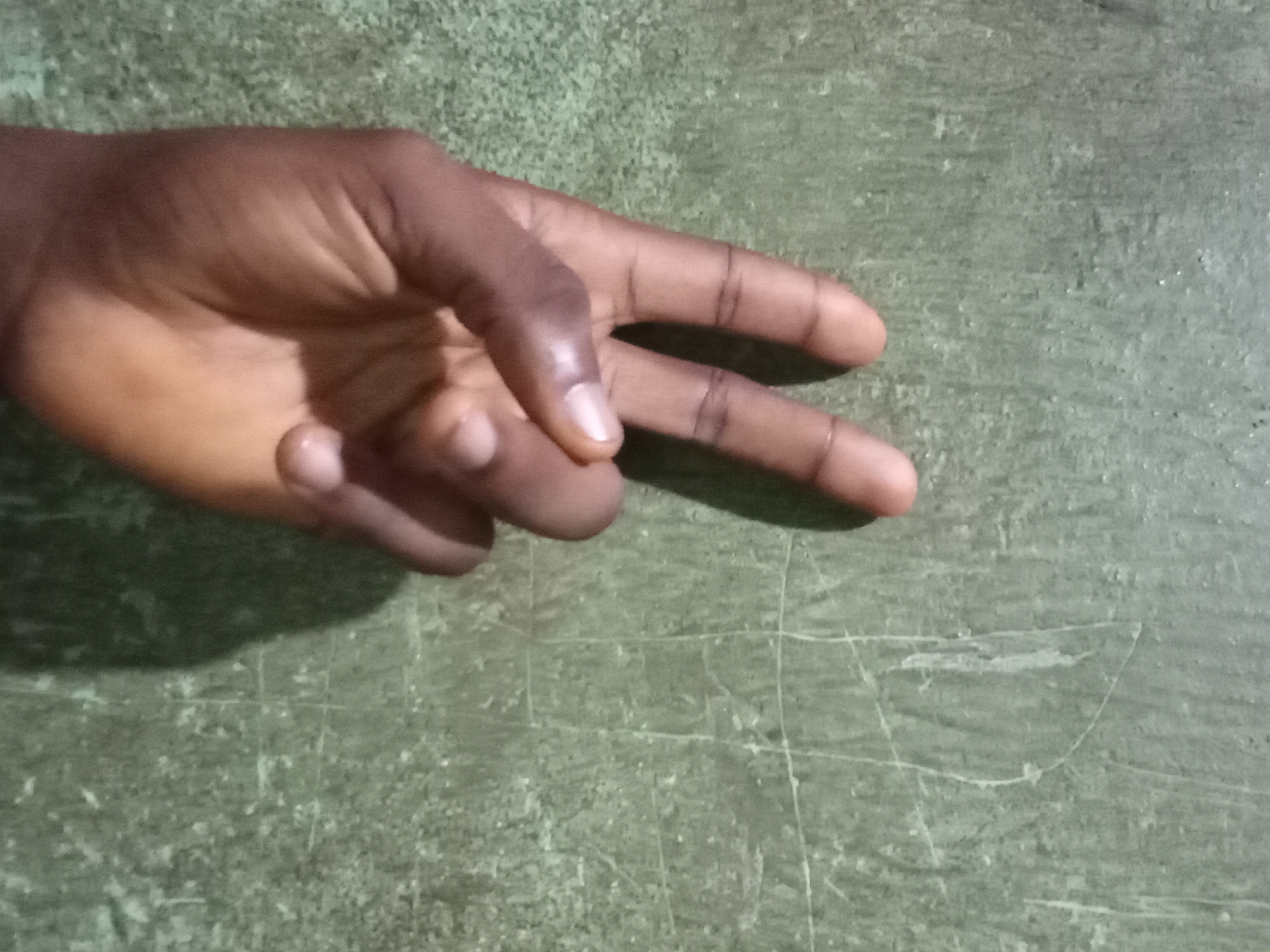 In Nkpor, Nigeria, a close-up shot captures a hand displaying the peace sign against a textured green wall. The wall exhibits signs of wear, with faint white scratches and a slightly mottled surface, suggesting it is an outdoor or weathered surface. The hand, belonging to an individual with darker skin tone, is positioned centrally in the frame. Two fingers, the index and middle finger, are extended upwards in a V-shape, while the thumb, ring finger, and pinky finger are curled inwards towards the palm. The lighting casts a soft shadow of the hand onto the wall, emphasizing the three-dimensional form of the hand and its gesture. The overall scene is simple and direct, focusing on the hand and its symbolic gesture against a utilitarian background.