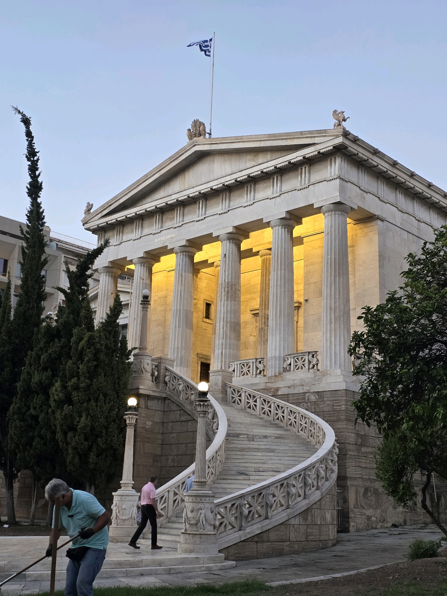 The image shows the National Library of Greece in Athens, a neoclassical building with a grand entrance featuring a sweeping marble staircase. The library is illuminated by warm, yellow light, highlighting its stately columns and architectural details. A Greek flag flies proudly from a flagpole on the roof.
In the foreground, a man with gray hair, wearing a light blue t-shirt and blue jeans, is raking the grass. He is focused on his task, holding a rake with both hands. Behind him, a man in a pink shirt and black pants walks up the staircase, while another person in a blue shirt is partially visible further up the steps.
To the left of the library, tall cypress trees stand against the pale blue sky. To the right, a leafy green tree partially obscures the building. The overall scene suggests an early evening setting, with the golden light creating a peaceful and majestic atmosphere.