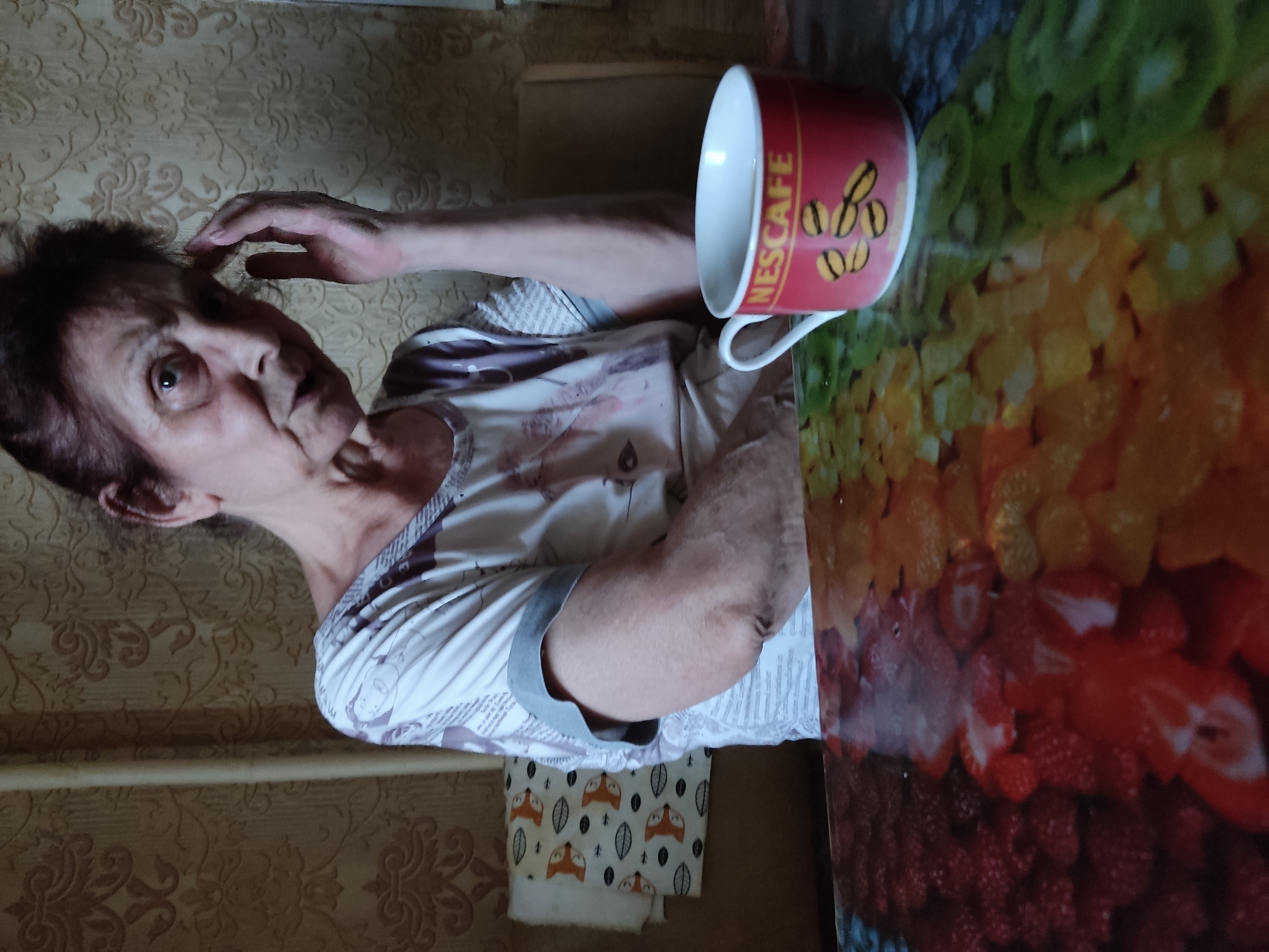 A medium shot captures an elderly woman with short, dark hair and fair skin, looking directly at the camera with a worried expression, her right hand touching her forehead. She is wearing a white t-shirt with a gray collar and a printed pattern. She is seated at a table with a colorful surface depicting sliced fruits. To her right, a red mug with the "NESCAFÉ" logo and an image of coffee beans sits on the table. In the background, a wall with floral wallpaper and a white towel with a fox print are visible. The scene appears to be indoors, possibly in a home setting.