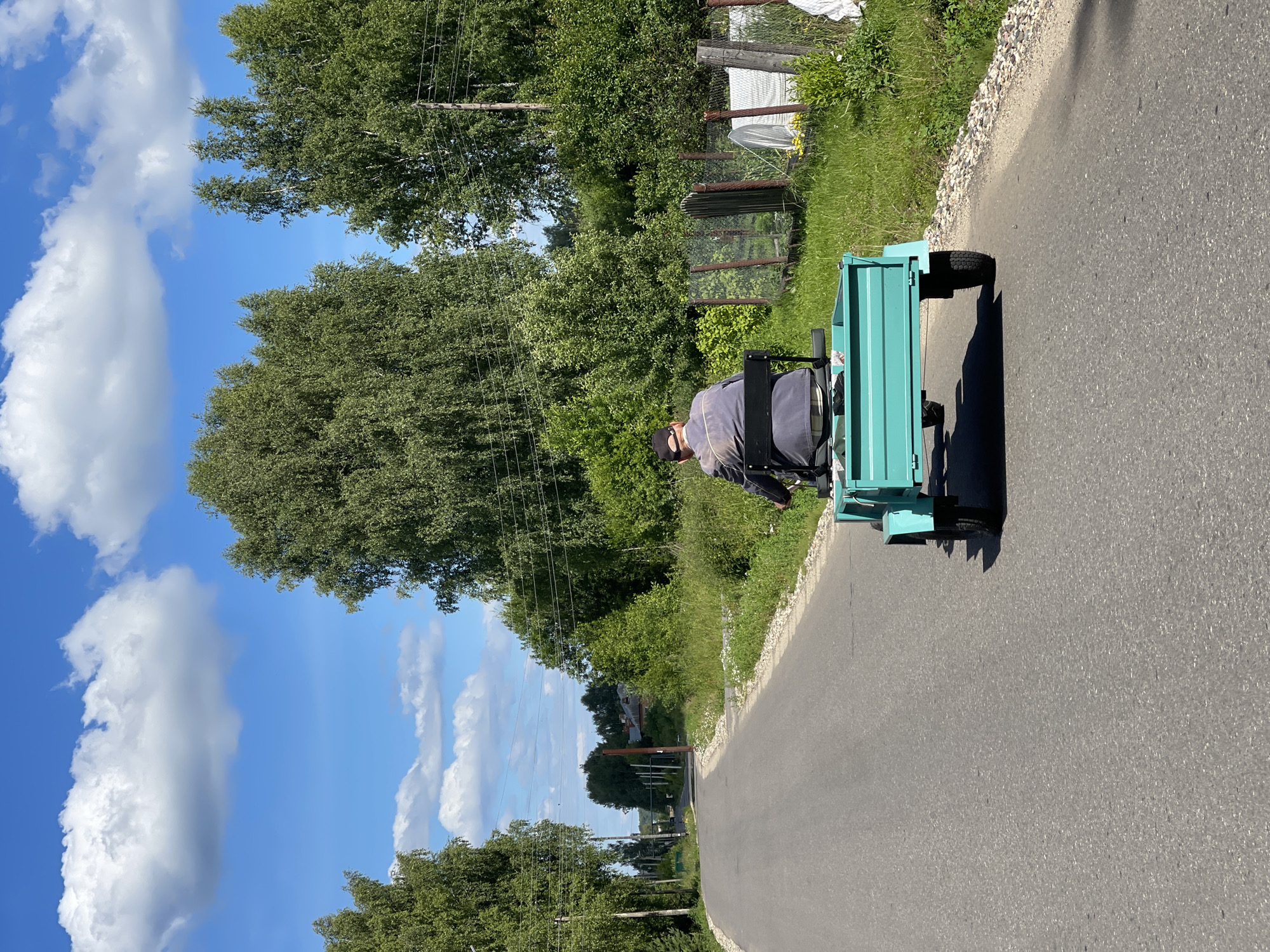 An individual wearing a dark jacket and a black cap is seated on a small, teal-colored motorized vehicle with a trailer attached. The vehicle is traveling on an asphalt road that curves to the left. The road is bordered by green grass and trees on both sides. In the background, there are more trees and some houses with red roofs. The sky is blue with fluffy white clouds. The overall scene depicts a rural or semi-rural environment on a clear day. The location is specified as the city of Заречный, Russia.