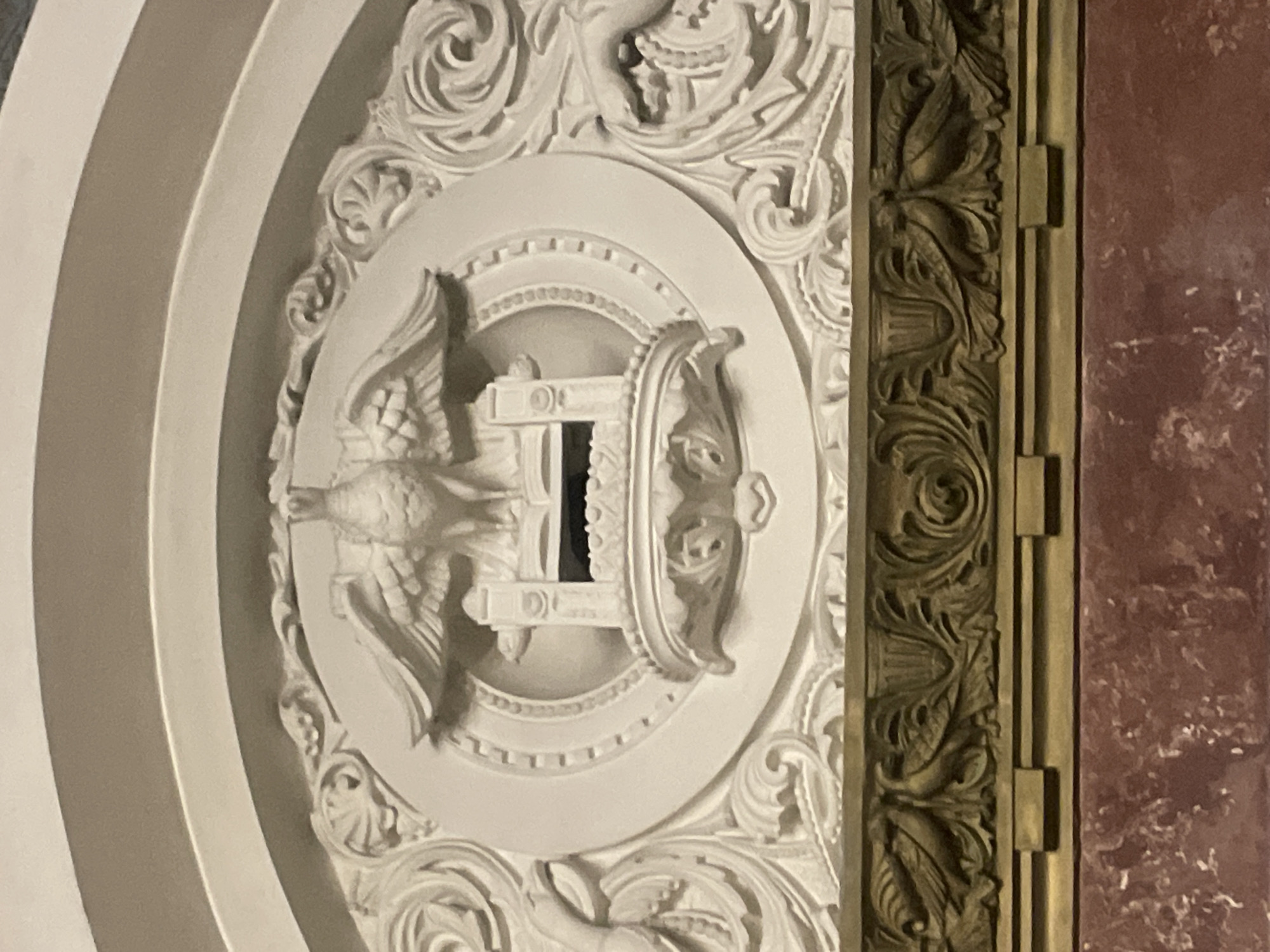 An ornate relief sculpture is shown, centrally featuring an eagle with outstretched wings above an open book resting on a pedestal. The sculpture is framed by decorative scrollwork and foliage. Below this white relief is a horizontal band of carved brass with foliate motifs and small bird-like figures. Beneath the brass band is a reddish-brown marble surface. The architectural style suggests a historical public building, possibly with classical or neoclassical influences. The image does not provide specific contextual information regarding its location in Kronstadt, Russia.