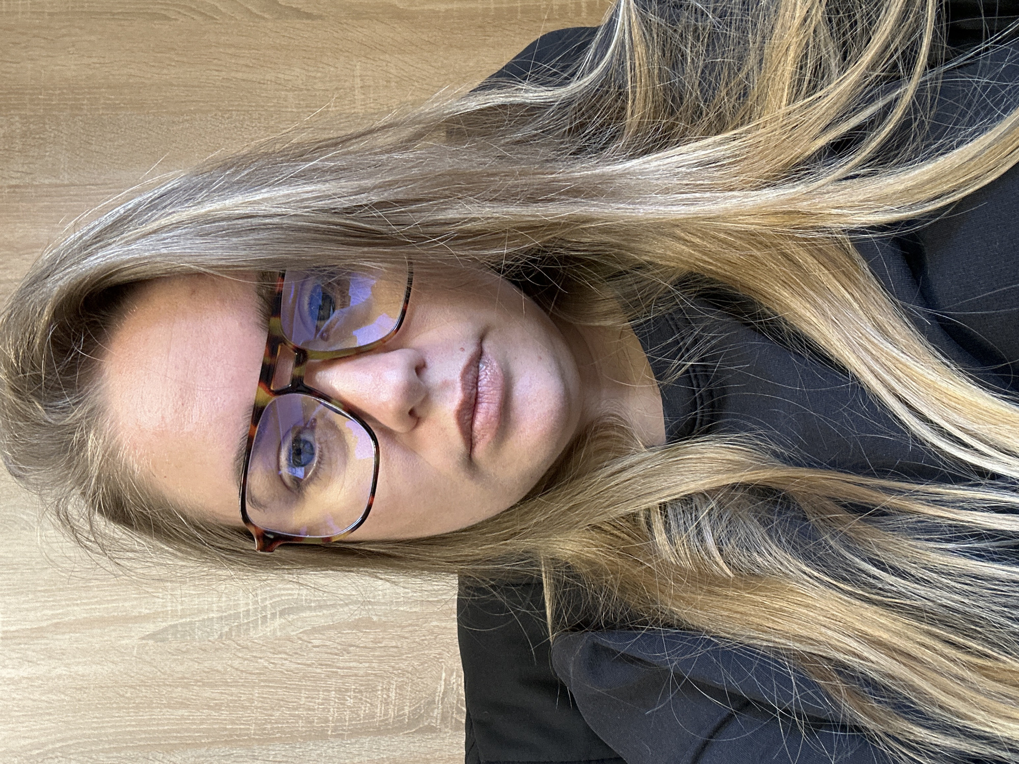 A woman with long, blonde hair and fair skin is wearing glasses with a brown tortoiseshell frame. She is wearing a black top or jacket. The background is a light brown surface resembling woodgrain. The image appears to be taken indoors with artificial lighting. There is no information in the image to suggest a location within the city of Ural'skoe, Russia.
