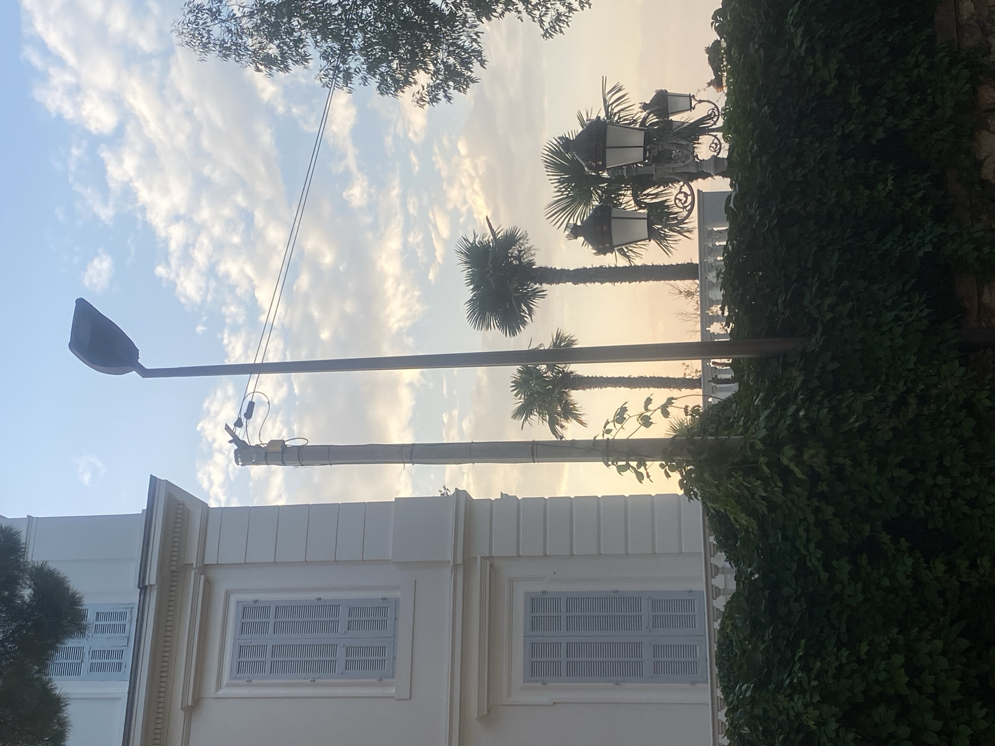 The image depicts a building with pale yellow walls and white shutters on the windows, situated behind a dense green hedge. The building appears to be a villa or a large house, possibly in Antibes, France, given the presence of palm trees which are common in the region. A modern street lamp is visible on the right side of the image, with an older, more ornate lamppost behind the hedge, suggesting a mix of architectural styles. The sky is a soft blue with scattered white clouds, indicating either early morning or late afternoon light. The overall scene suggests a tranquil, Mediterranean setting.
