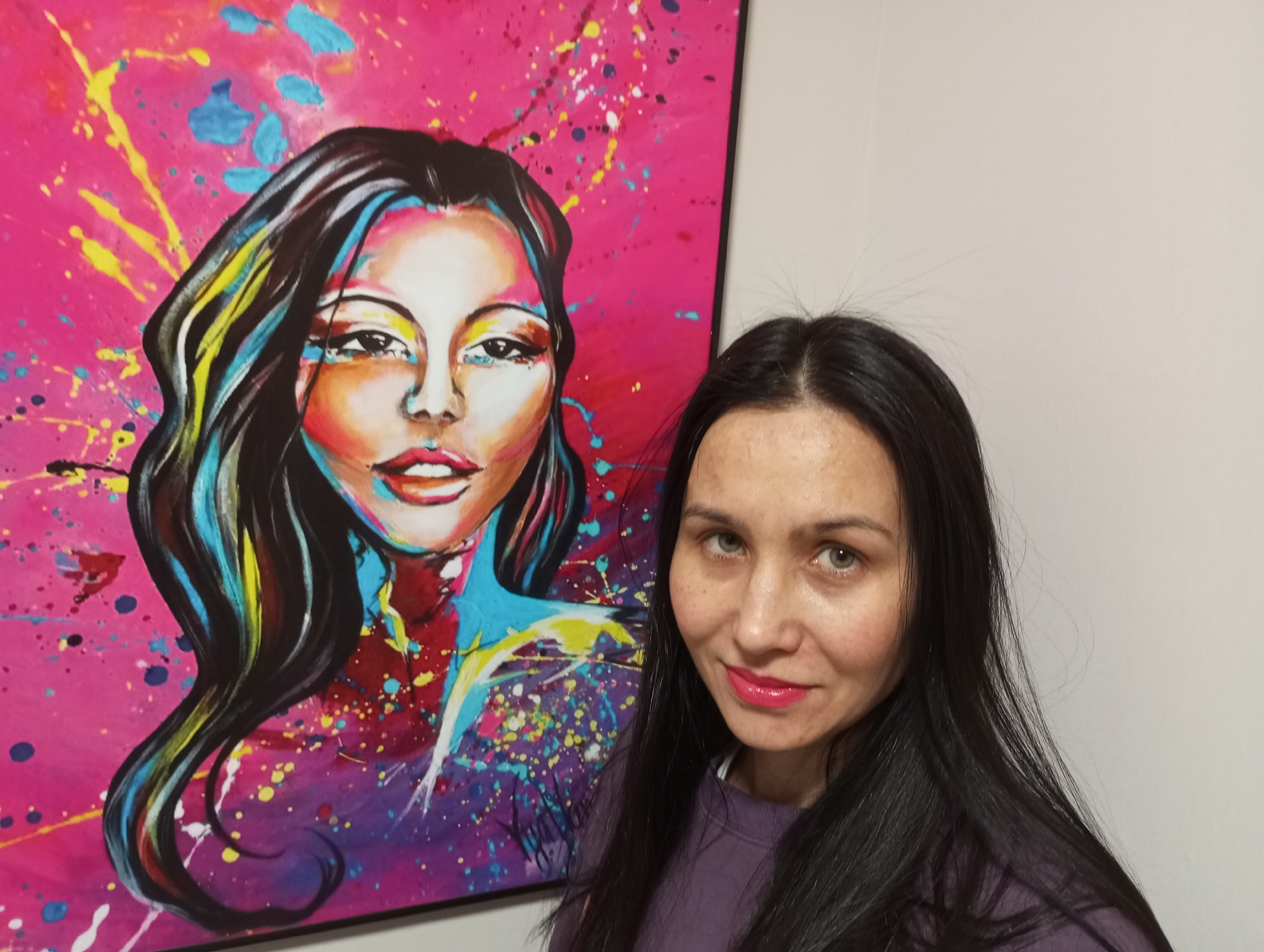 A woman with long dark hair stands indoors next to a framed painting. The painting depicts a woman with dark hair against a pink background with splashes of yellow, blue, and purple paint. The woman in the photo is wearing a purple sweater and lipstick. The wall is light gray. The image was likely taken in a private residence. No evidence suggests a location in Pervomaysky, Russia.