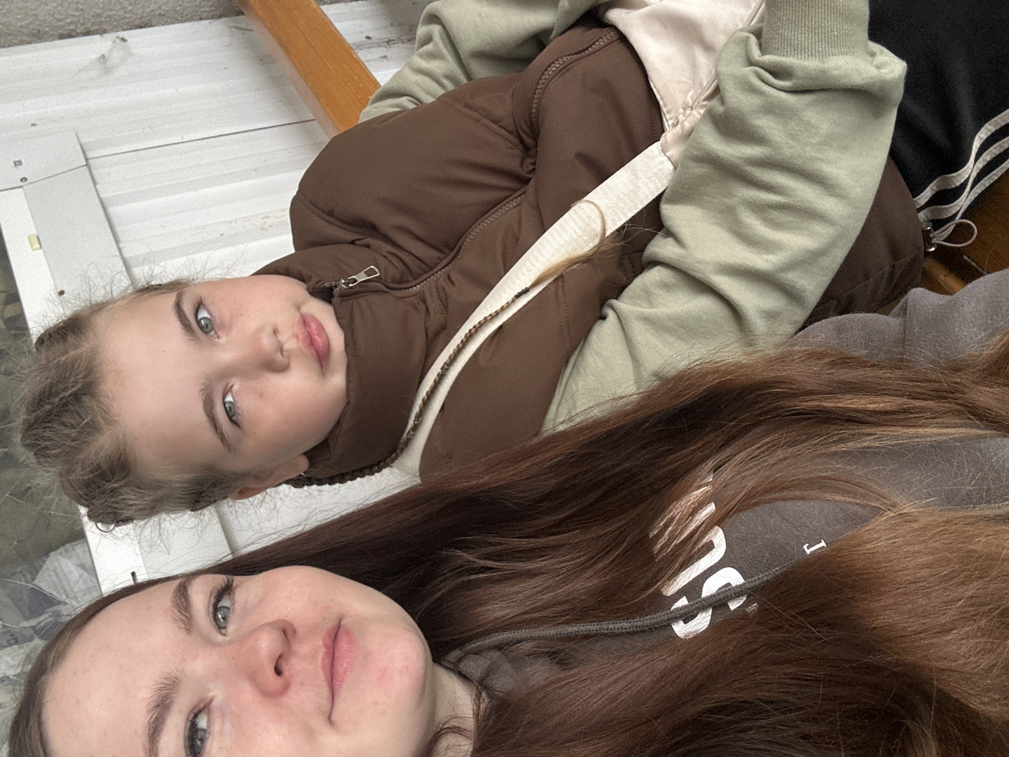 Two individuals are positioned in a close-up indoor or sheltered outdoor setting within the city of Chaikovsky, Russia. The person on the left, a young adult female, has long reddish-brown hair and light eyes, and is wearing a dark grey hooded sweatshirt with partial white lettering visible ("SU" or "SLI", "I", "Γ"). She is positioned slightly forward and to the left, looking directly towards the camera. The second individual, a child, is positioned to the right and slightly behind, with light hair styled in braids or small buns and light eyes, exhibiting a slightly pouting expression. She is wearing a brown puffer vest over a light olive green long-sleeved top, with a beige or white strap diagonally across her chest, possibly from a bag. Her lower right side reveals black pants with white stripes. The background features white vertical paneling, possibly a wall or siding, with some minor discoloration and a small yellow fixture. To the right, a wooden railing with an orange-brown stain is partially visible. The lighting is even and soft.