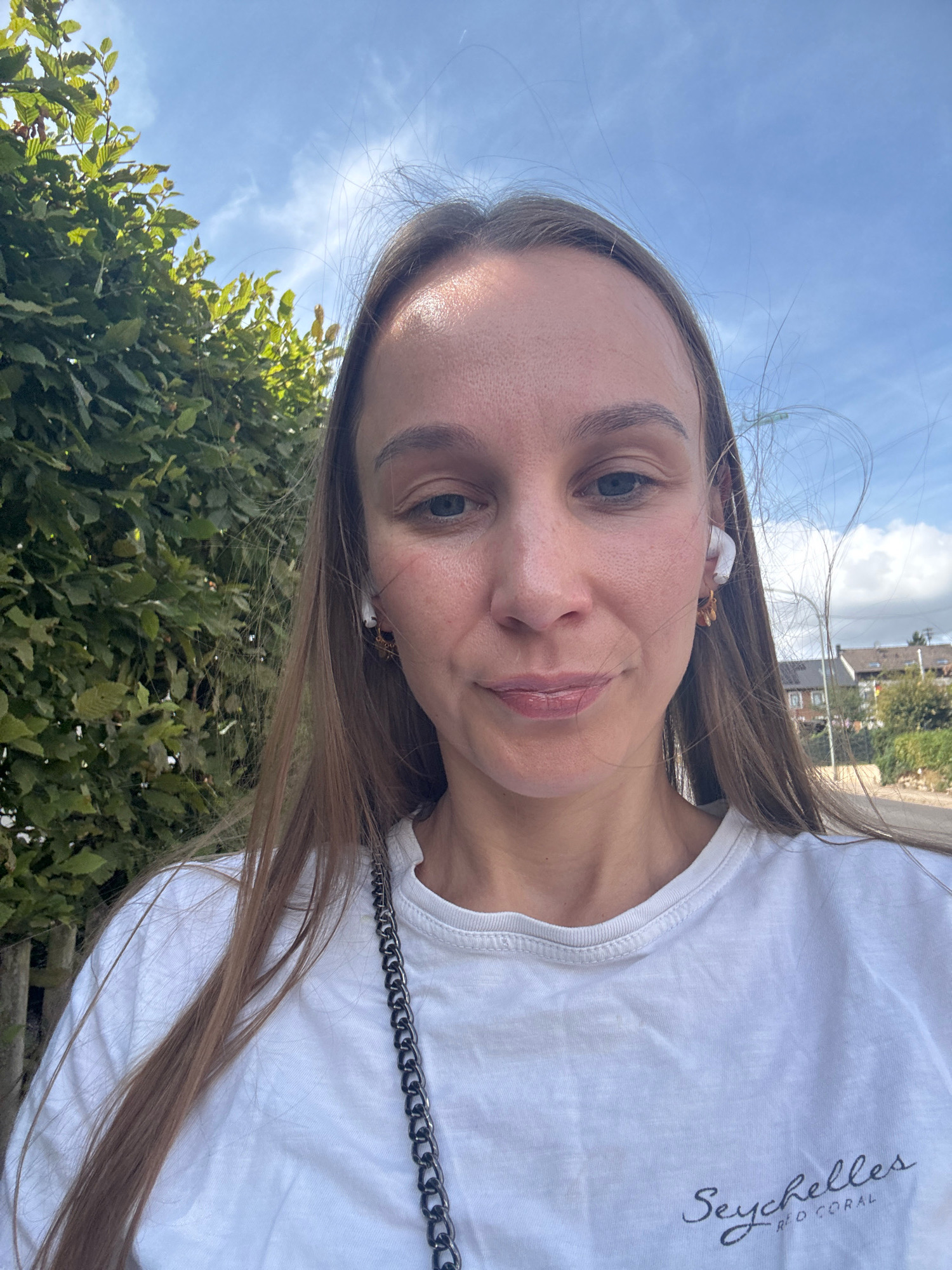 A woman is taking a selfie outdoors in what appears to be a residential area in Überherrn, Germany. She has long, light brown hair and is wearing a white t-shirt with the text "Seychelles RED CORAL" printed on it. She has white wireless earbuds in her ears and is wearing small gold earrings. Her left shoulder is visible, and a dark chain strap is draped across her t-shirt. Behind her, there is a dense green bush on the left side of the frame and a row of houses with tiled roofs in the background on the right. The sky is blue with some scattered clouds. The lighting suggests it is daytime.