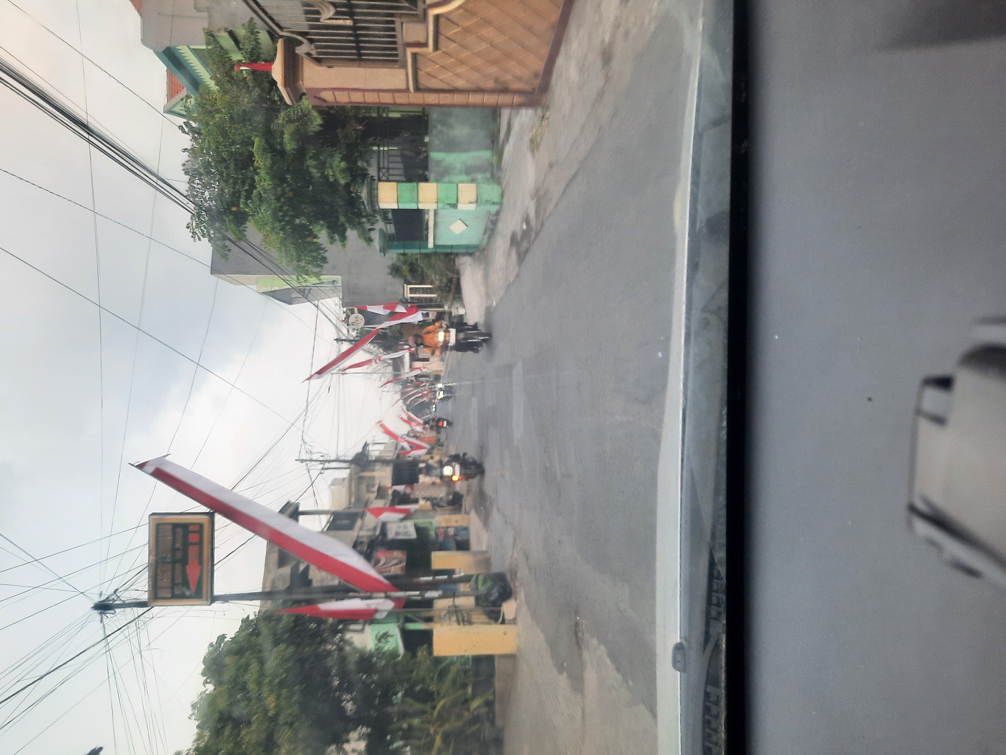 The image depicts a street scene in Banjarkemantren, Indonesia, adorned with numerous Indonesian flags. The flags, predominantly red and white, are strung along the street and displayed on poles, suggesting a patriotic celebration or event. Several motorcycles are visible on the asphalt road, with individuals riding them, indicating ongoing traffic. Buildings and trees line the sides of the street, and overhead power lines are prominent. The sky is overcast. The perspective is from inside a vehicle, looking forward through the windshield.