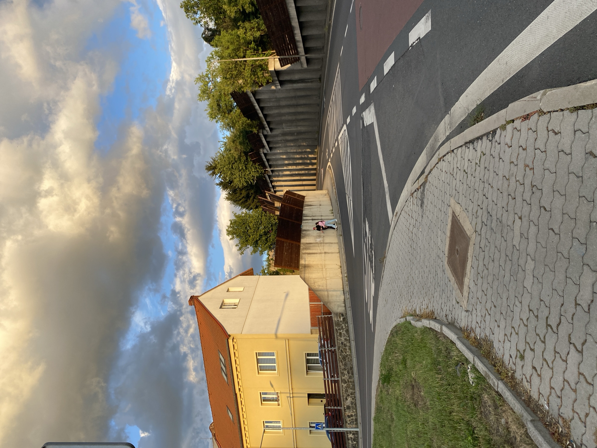 The image depicts a street scene in Bílina, Czechia, likely during the late afternoon or early evening, judging by the lighting and cloud formations. A yellow and white house with an orange roof is visible on the left side of the frame. Adjacent to the house is a concrete structure with wooden fencing.
The road features a pedestrian crossing with white stripes. A paved sidewalk runs alongside the road, curving away from the viewer. Green grass borders the sidewalk. On the right side of the road, a tall concrete retaining wall with wooden elements separates the road from a tree-covered embankment. The sky is filled with a dramatic mix of dark and light clouds, with patches of blue sky visible.
Two individuals, one wearing a pink shirt and the other dark clothing, are walking on the sidewalk near the house, moving away from the viewer. Two cars are parked in front of the house, behind a wooden fence. A blue and white pedestrian crossing sign is visible near the parked cars. The overall impression is an urban setting with residential buildings and infrastructure, set against a dynamic sky.