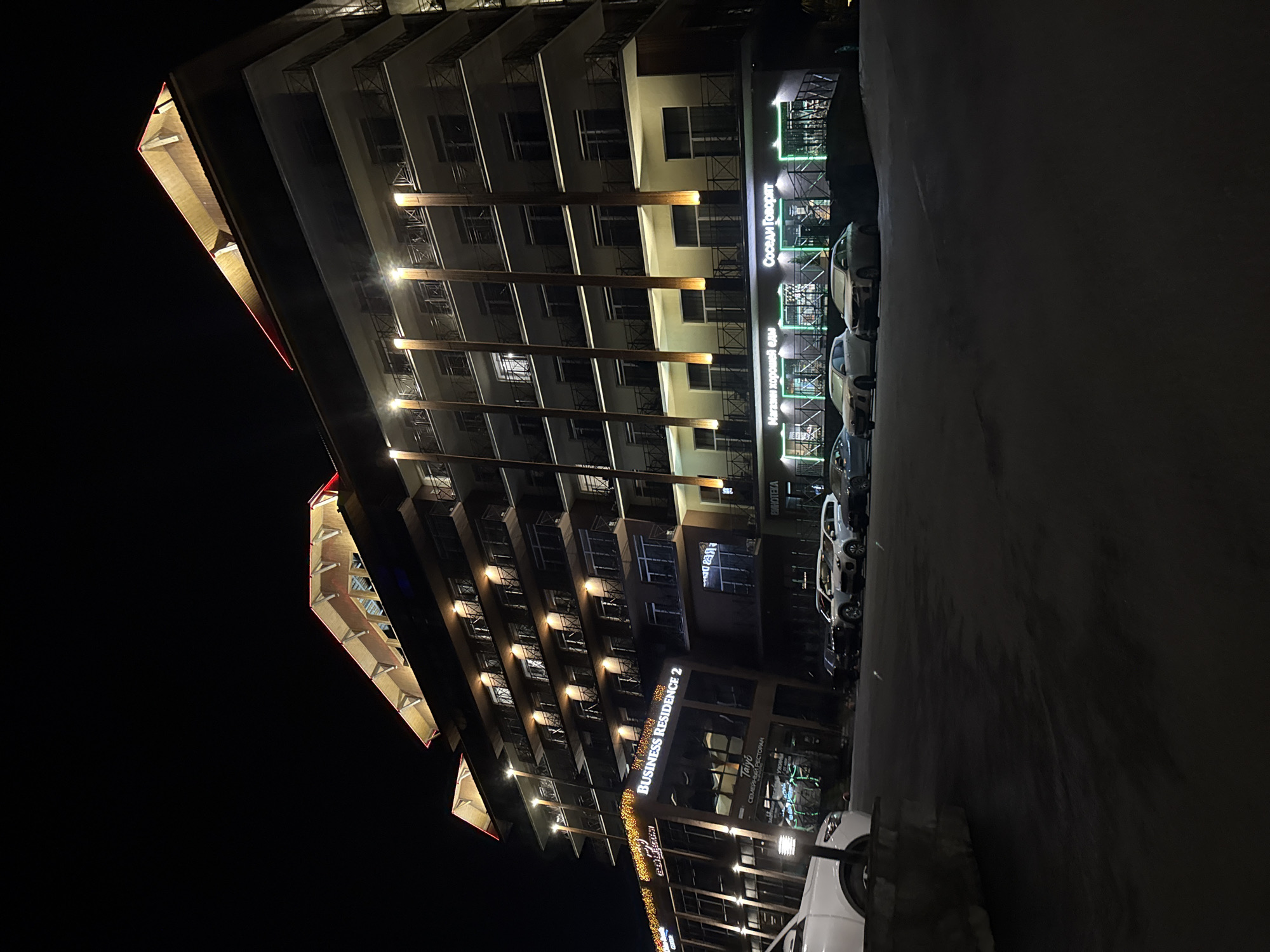 An exterior nighttime shot showcases the "Business Residence 2" building in Syrostan, Russia. The multi-story structure features numerous balconies with black railings and is illuminated by warm yellow lights along its facade and red accent lighting on its roof. A white SUV is parked in the foreground on the left, facing right. Several other cars, including white and dark sedans and SUVs, are parked in front of the building's ground-floor businesses, which are identifiable by green neon signage. The ground level has large glass windows displaying products and interiors. The area appears to be a parking lot with a dark asphalt surface, and some low landscaping is visible. The sky is dark, indicating a nighttime scene.