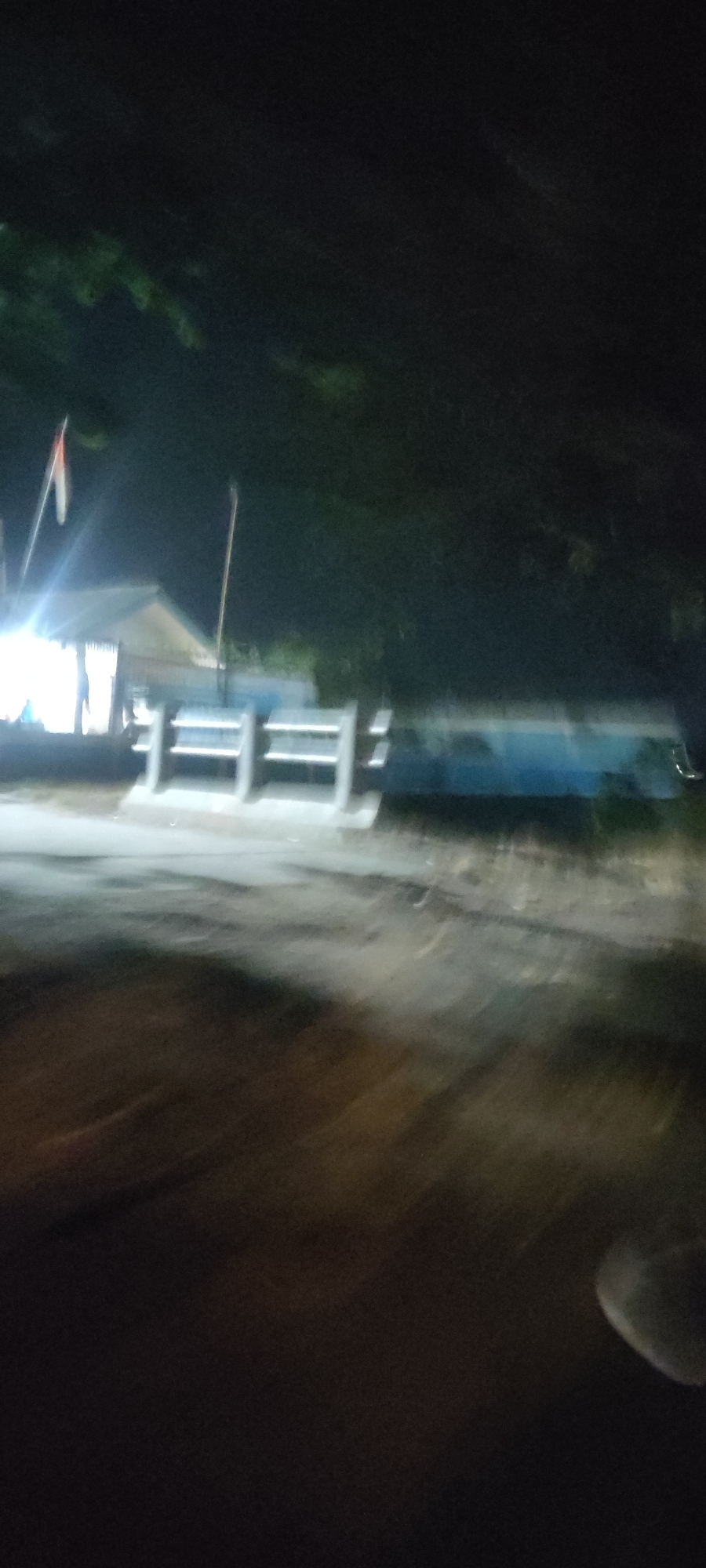 This is a blurry, nighttime photograph taken at the Babakangebang, Indonesia location. In the background, a building with a light-colored roof is visible, with a flag flying above it. In front of the building, there are several concrete pillars with horizontal bars, possibly part of a fence or barrier. To the right of this structure, a long, blue object, possibly a vehicle or a building facade, extends across the frame. The foreground is dominated by a blurred, dark surface, likely a road, with some lighter patches suggesting wetness or reflections. The overall impression is one of a dimly lit, possibly rural or semi-urban setting at night.