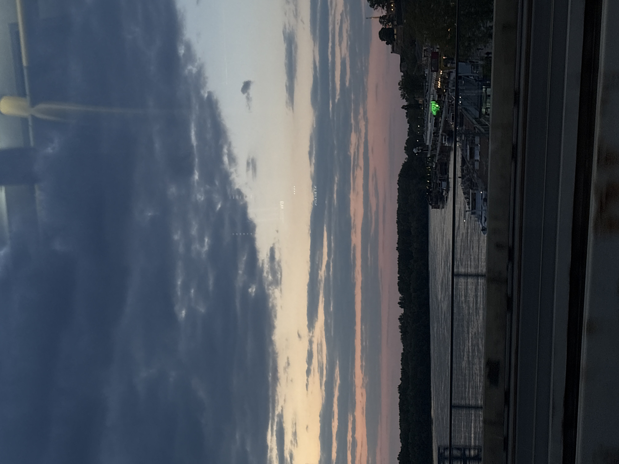 The image captures a twilight scene over a wide body of water, likely a river, viewed from an elevated position such as a bridge or through a window of a vehicle. The sky dominates the upper portion, featuring dark grey and blue clouds on the left, gradually transitioning to lighter shades of blue, grey, and faint pinks and oranges towards the right horizon, indicative of dusk.
The water below reflects the subdued light of the sky, showing ripples and shimmer. On the far bank, a dark, tree-lined landmass stretches across the horizon. Along this shoreline, various industrial or dock-like structures are visible, illuminated by scattered artificial lights, including a prominent green light. Several boats or barges appear to be moored near these structures.
In the foreground on the right, the strong vertical and horizontal lines of a bridge's support structure or railings frame the scene. The left side of the image shows reflections and glare on what appears to be a glass surface, suggesting the photograph was taken from inside a vehicle. Faint, mirrored text, possibly "WB" and "ZJO.012", can be observed within these reflections.
The overall atmosphere is calm and contemplative, reflecting the transition from day to night. Given the provided context, this scene depicts a view over a river in Belgrade, Serbia, likely one of the major rivers like the Sava or Danube.