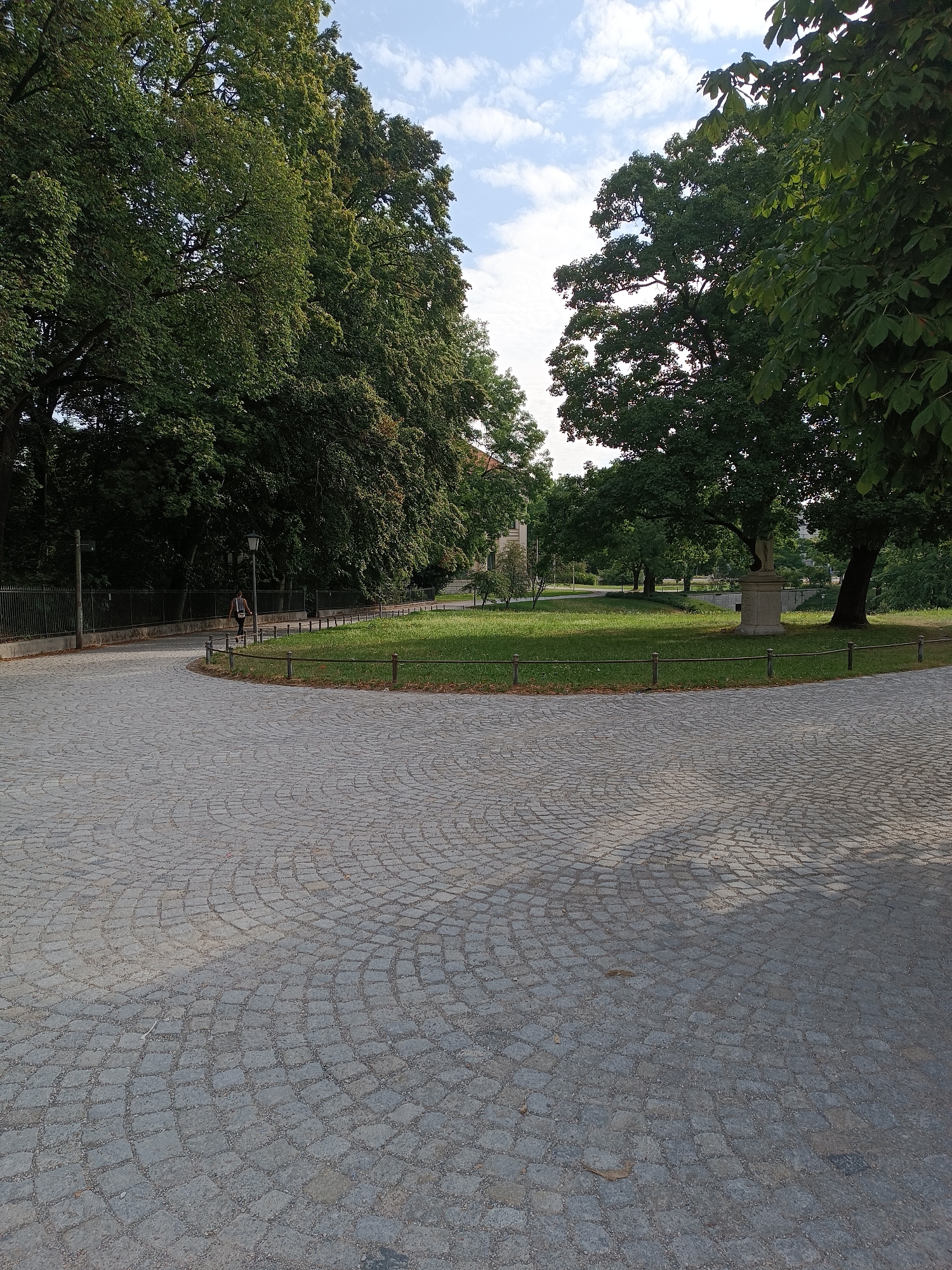 The image depicts a paved pathway made of cobblestones that curves to the left. The pathway is bordered by a grassy area on the right and a line of trees and a fence on the left. A single person is visible walking on the pathway, wearing dark clothing. Beyond the grassy area, there are more trees and a statue on a pedestal. The sky above is partly cloudy. The location is in Munich, Germany.