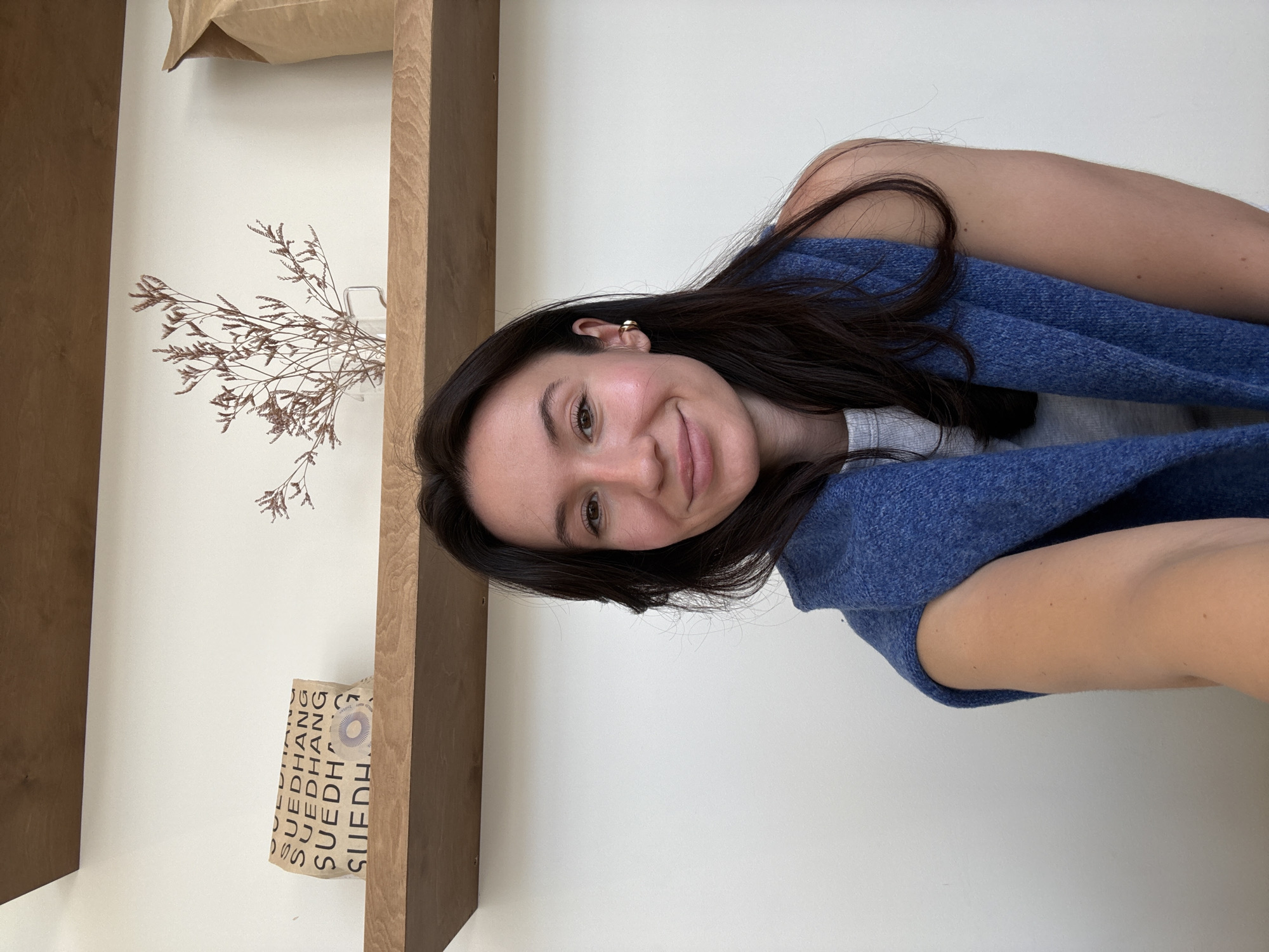 A young woman with dark brown hair is centered in the frame. She is smiling gently at the camera. She appears to be indoors, positioned in front of a white wall with a wooden shelf to the left. On the shelf, there is a paper bag with a logo or writing that includes "SUEDHANG" and a small dried flower arrangement. The woman is wearing a blue sweater. The location is potentially Tel-Aviv, Israel, but there is no definitive visual evidence confirming the city.