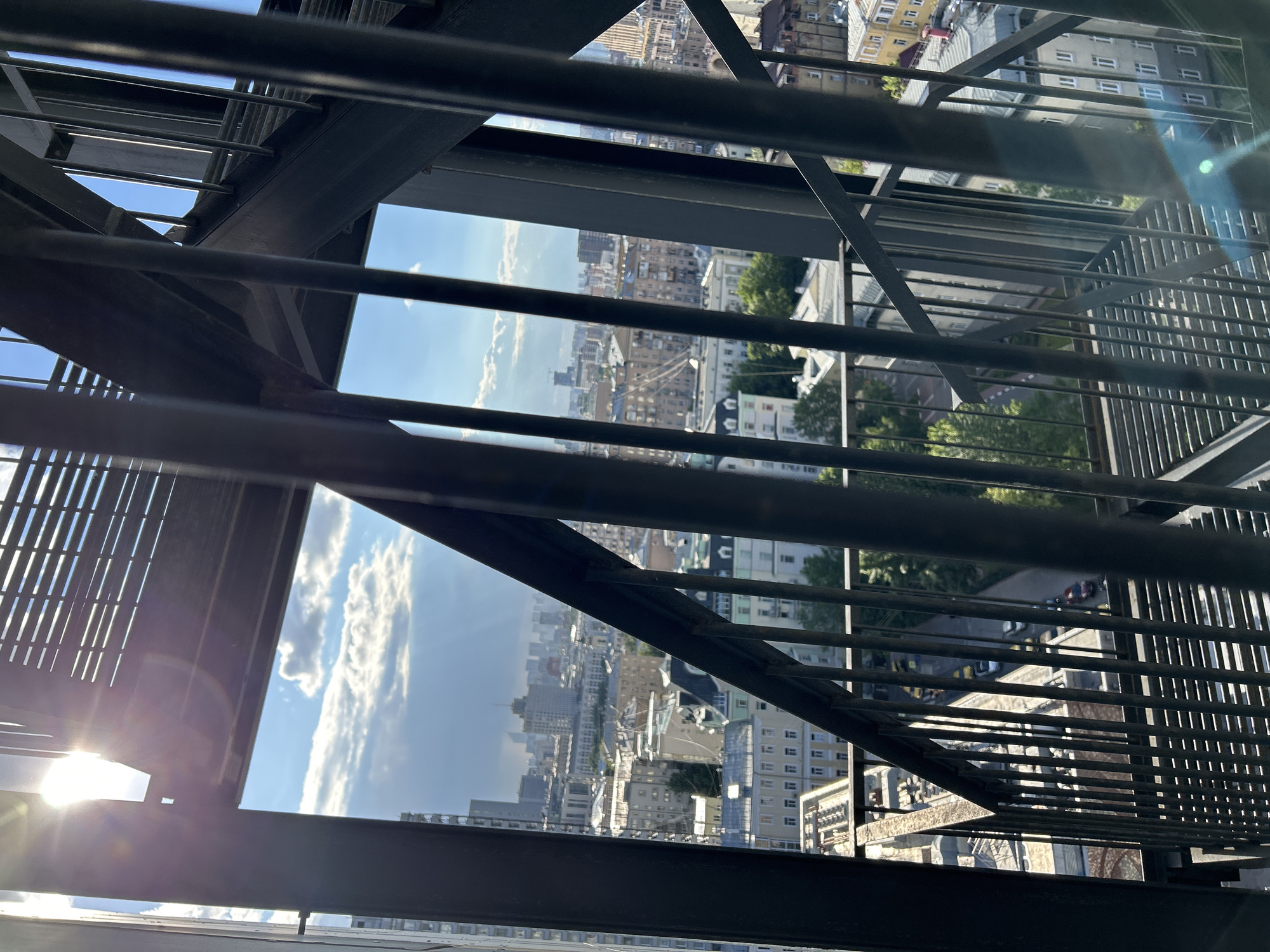 The image is taken from an elevated perspective, looking out over a cityscape through a metal structure, possibly a fire escape or scaffolding. The city of Unknown, Unknown, is partially obscured by the metal framework. Buildings of varying heights and architectural styles fill the background. The sky is visible in patches, with blue and white clouds. The sun is visible in the lower left corner.