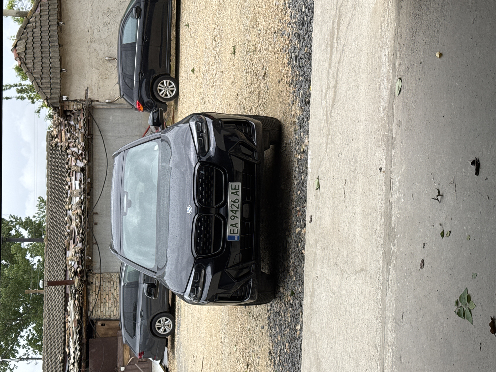 The image shows the front of a black BMW X3 parked on a gravel surface. The car has license plates that read "EA 9426 AE". In the background, there is a building with a damaged tiled roof and two other cars parked. The scene appears to be in a residential or industrial area. The weather is overcast.