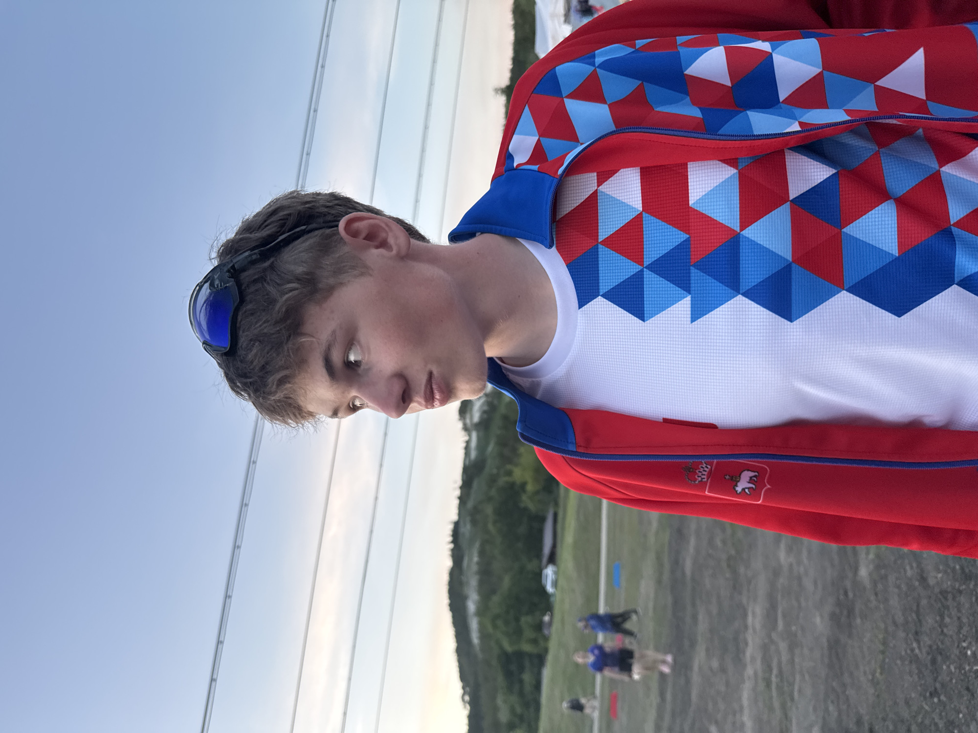 A young man stands outdoors in a red, white, and blue patterned jacket and white t-shirt, with sunglasses perched on his head. He gazes downward. In the background, a grassy field leads to a wooded area. Power lines cross the sky. Several figures are visible on the field. The logo of Khvalynsk is visible on the jacket, suggesting the location is the city of Khvalynsk, Russia.
