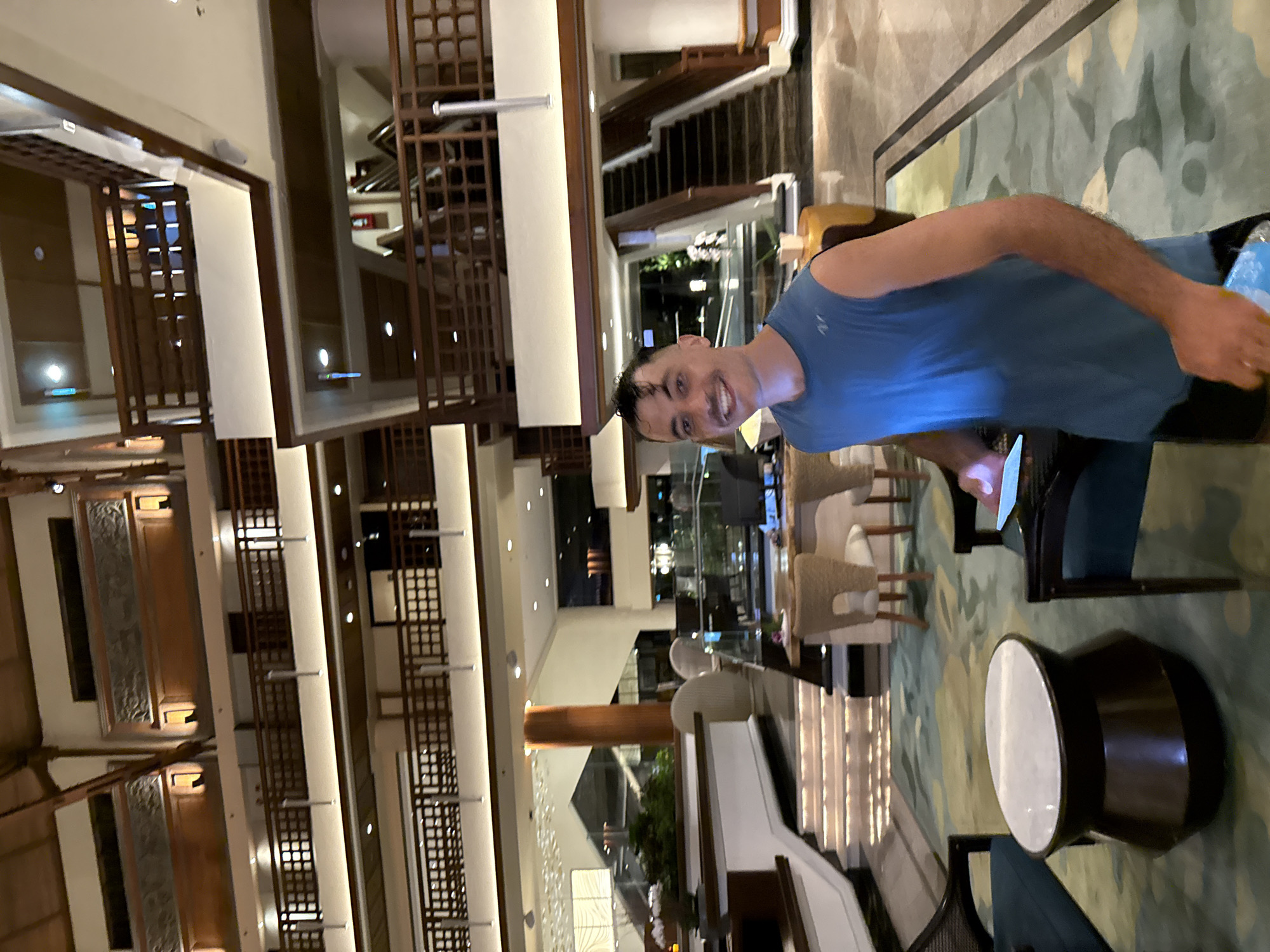 The image shows a man in a blue tank top smiling at the camera while sitting in what appears to be a hotel lobby. The lobby has multiple levels with ornate wooden railings. The interior design incorporates neutral tones, modern furniture, and decorative lighting. Through large windows, there are glimpses of outdoor landscaping. The location is identified as Unknown, Indonesia.