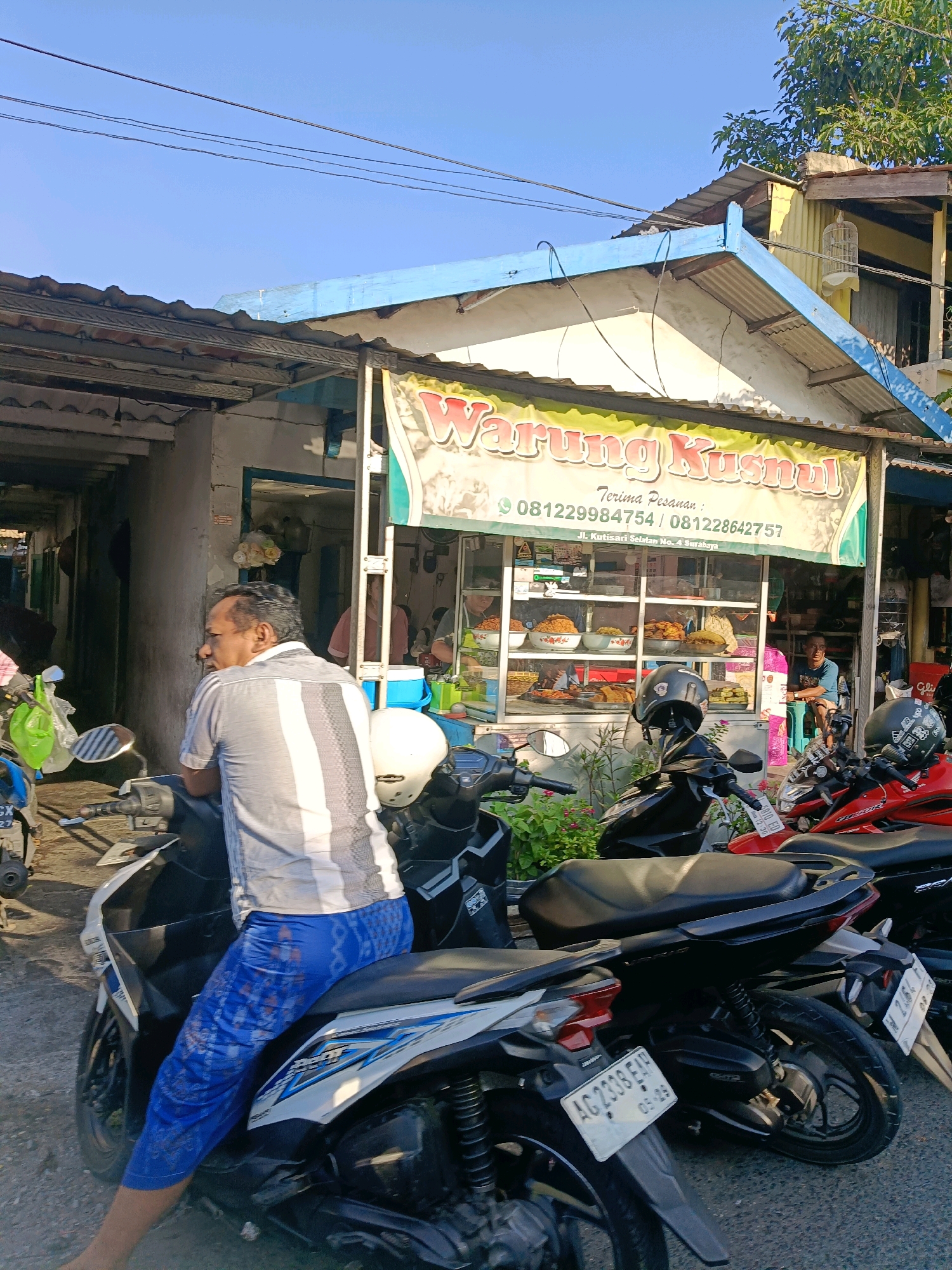 The image depicts a street scene in Surabaya, Indonesia, in front of a food stall identified by a banner as "Warung Kusnul." The stall appears to be open, with food items displayed in glass cabinets. Several motorcycles are parked in front of the stall, suggesting it's a popular spot. One man, wearing a gray and white striped shirt and blue patterned pants, is seated on a white motorcycle, facing away from the camera. Another man, wearing a blue t-shirt, is sitting on a stool inside the stall. The environment is a typical urban street with buildings and overhead wires visible. The lighting suggests it is daytime, likely morning or late afternoon due to the sun's angle.
