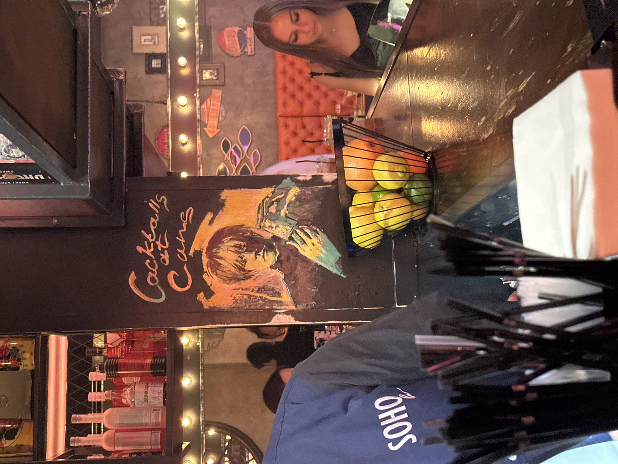 The image depicts a bar scene with a focus on the bar counter and the area behind it. In the foreground, a person wearing a blue shirt with "SOHO" printed on it is partially visible. A black metal basket filled with citrus fruits such as oranges, lemons, and limes sits on the wooden bar counter. Behind the counter, a woman with long dark hair is seated, wearing a black top, and looking down. The background features a wall with decorative elements, including framed pictures and neon lights. To the left, shelves are stocked with various bottles, some illuminated by pink neon lights. A hand-painted sign with the text "Cocktails at Cane" is prominently displayed on a dark surface. The overall environment suggests an indoor bar or lounge.