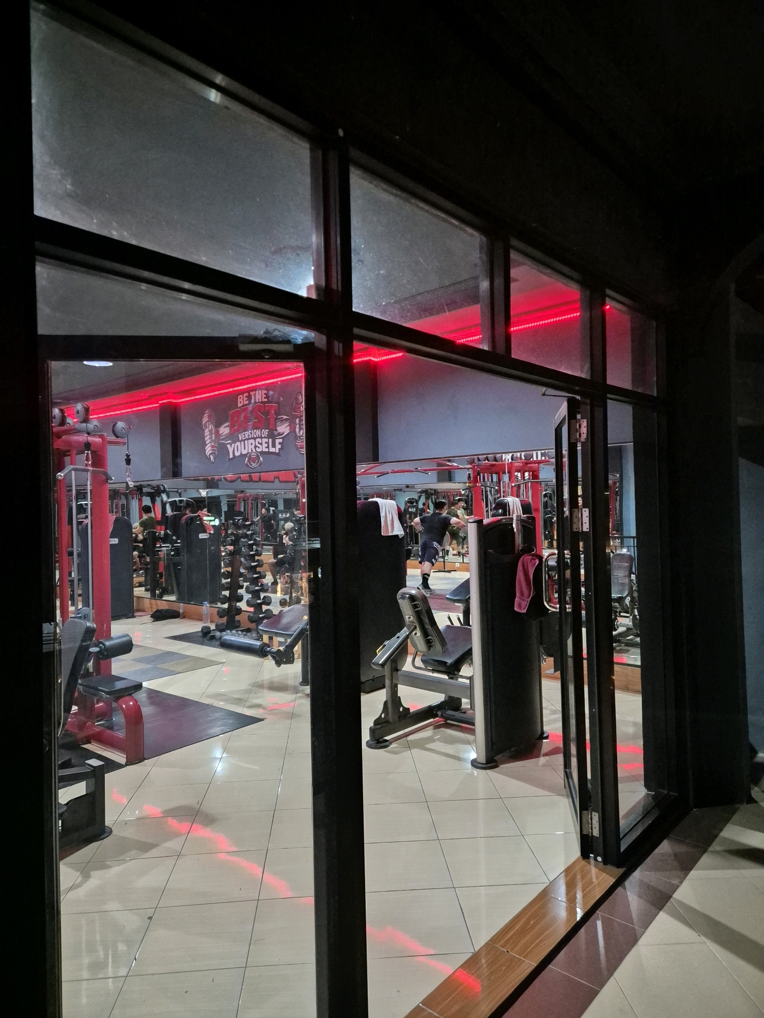The image shows the interior of a gym in Bontang, Indonesia, viewed from the outside through large glass doors. The gym is illuminated with red LED lighting, creating a vibrant atmosphere. Various exercise machines, including weight machines and dumbbell racks, are visible throughout the space. Several individuals are engaged in workouts, some using equipment and others in the process of exercising. The floor is tiled, and there is a motivational poster on the wall that reads "BE THE BEST VERSION OF YOURSELF." The time appears to be evening or night, given the darkness outside and the internal lighting.