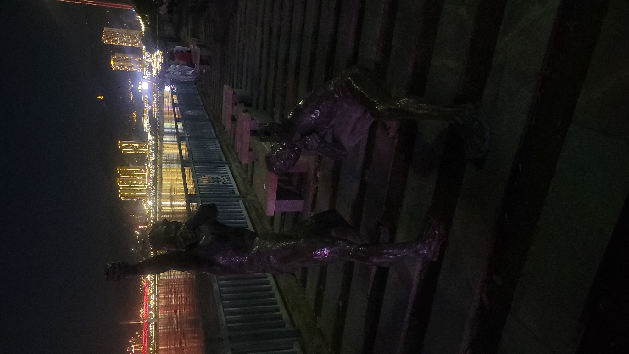At night in Nan'an District, China, two bronze statues stand on a walkway along a body of water. The statue in the foreground shows a man raising his fist in the air, while the statue in the background shows a man kneeling. The walkway is made of wood and concrete, and there are lights reflecting off the water in the background. Buildings illuminated with lights can be seen across the water. People are walking on the walkway in the distance.