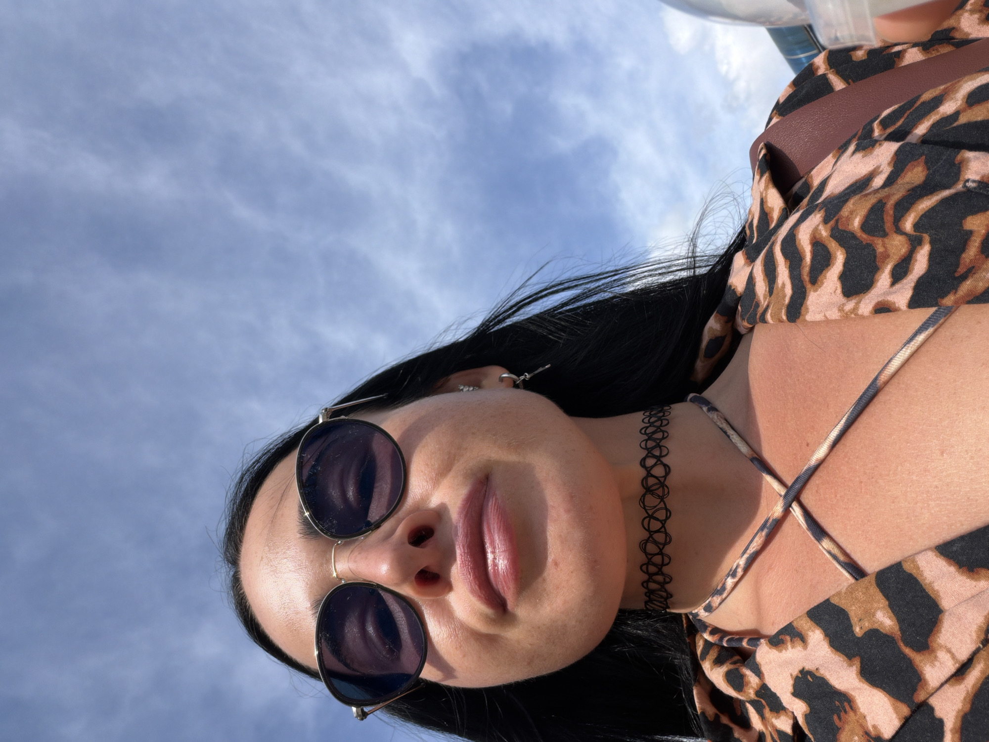 A close-up, low-angle selfie captures a woman with long, dark hair and fair skin, wearing large, round sunglasses, a leopard-print top with a criss-cross neckline, and a black choker necklace. She is looking upwards towards a partly cloudy blue sky, suggesting an outdoor setting, possibly in Sochi, Russia, given the bright daylight and sky conditions. A hint of a brown bag strap is visible on her shoulder.