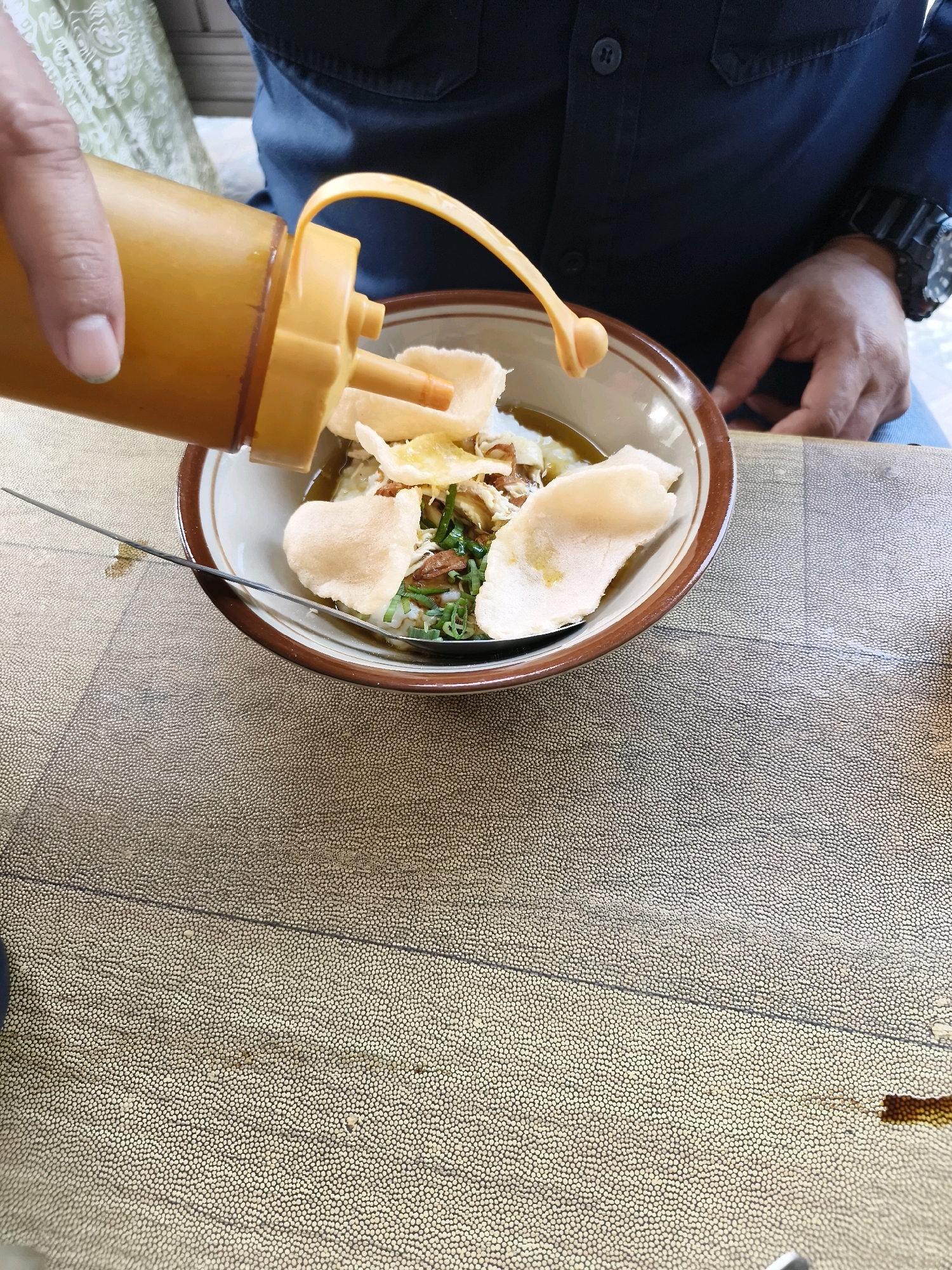 A close-up shot shows a person in a dark blue shirt with a watch on their left wrist pouring a sauce from a yellow squeeze bottle into a bowl of food. The food appears to be a savory dish, possibly congee or rice porridge, topped with crackers, shredded chicken, and green onions. A spoon rests in the bowl. The scene is set on a textured, light-colored table. The background shows a person's arm and shoulder, and a green patterned fabric. The lighting is natural and bright. The location is the city of Salira, Indonesia.