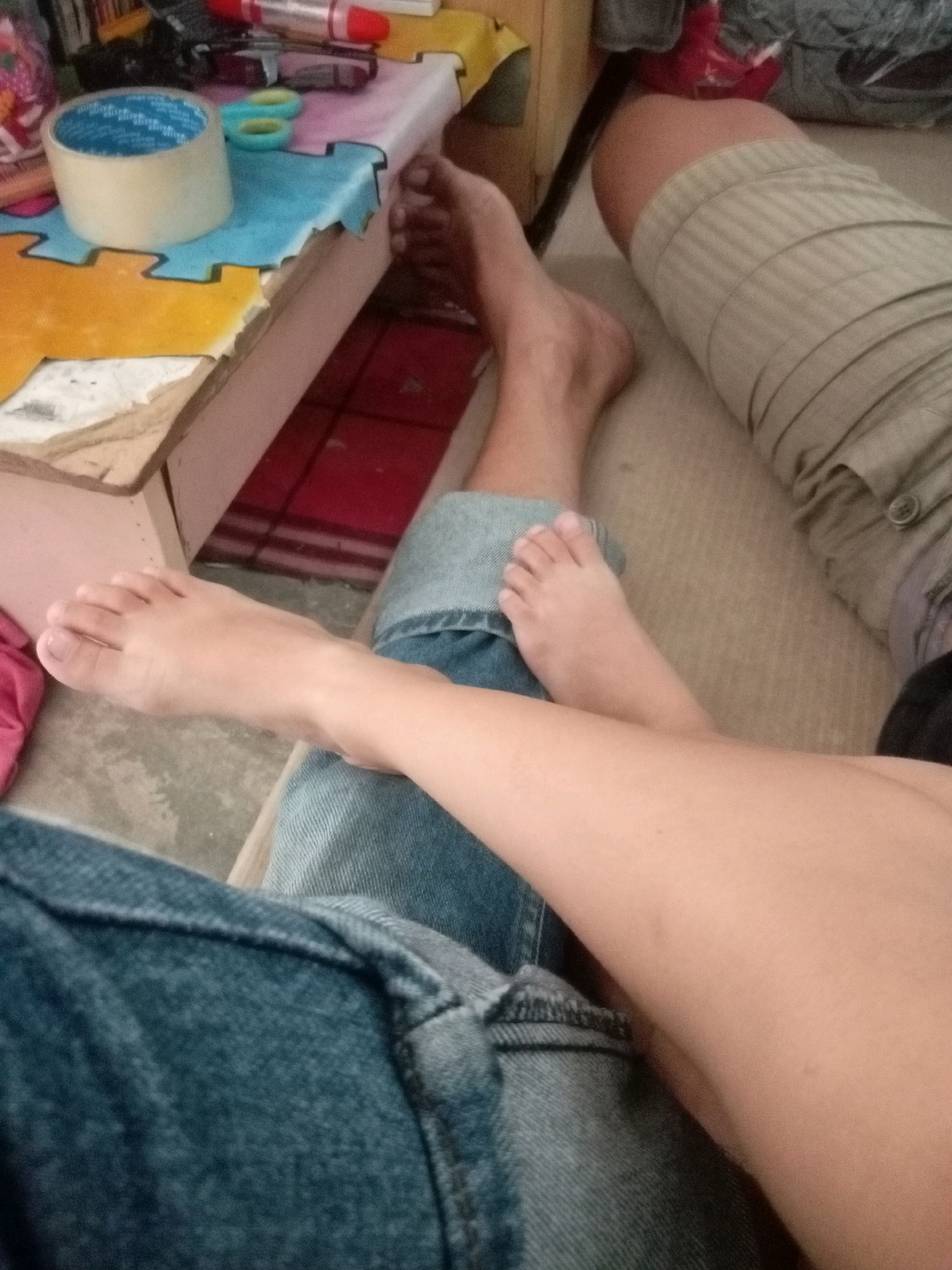 The image displays a close-up view of two individuals' legs and feet, suggesting a casual, indoor setting. One person's feet are bare and resting on what appears to be a beige carpet or upholstery. This person is wearing blue denim jeans. Adjacent to them, another individual's legs are visible, one clad in khaki-colored pants with a ribbed texture and a button detail, and the other leg is bare and crossed over the denim-clad leg. A small, bare foot is also visible, positioned between the two individuals' legs.
In the background, a low table is partially visible, holding various items including a roll of clear tape, a pair of light blue scissors, and other unidentifiable objects. The overall lighting suggests an indoor environment, likely a home, given the presence of personal items and the relaxed posture of the individuals. The image does not contain any discernible elements that would allow for specific location identification within Batam, Indonesia, or any other city.
