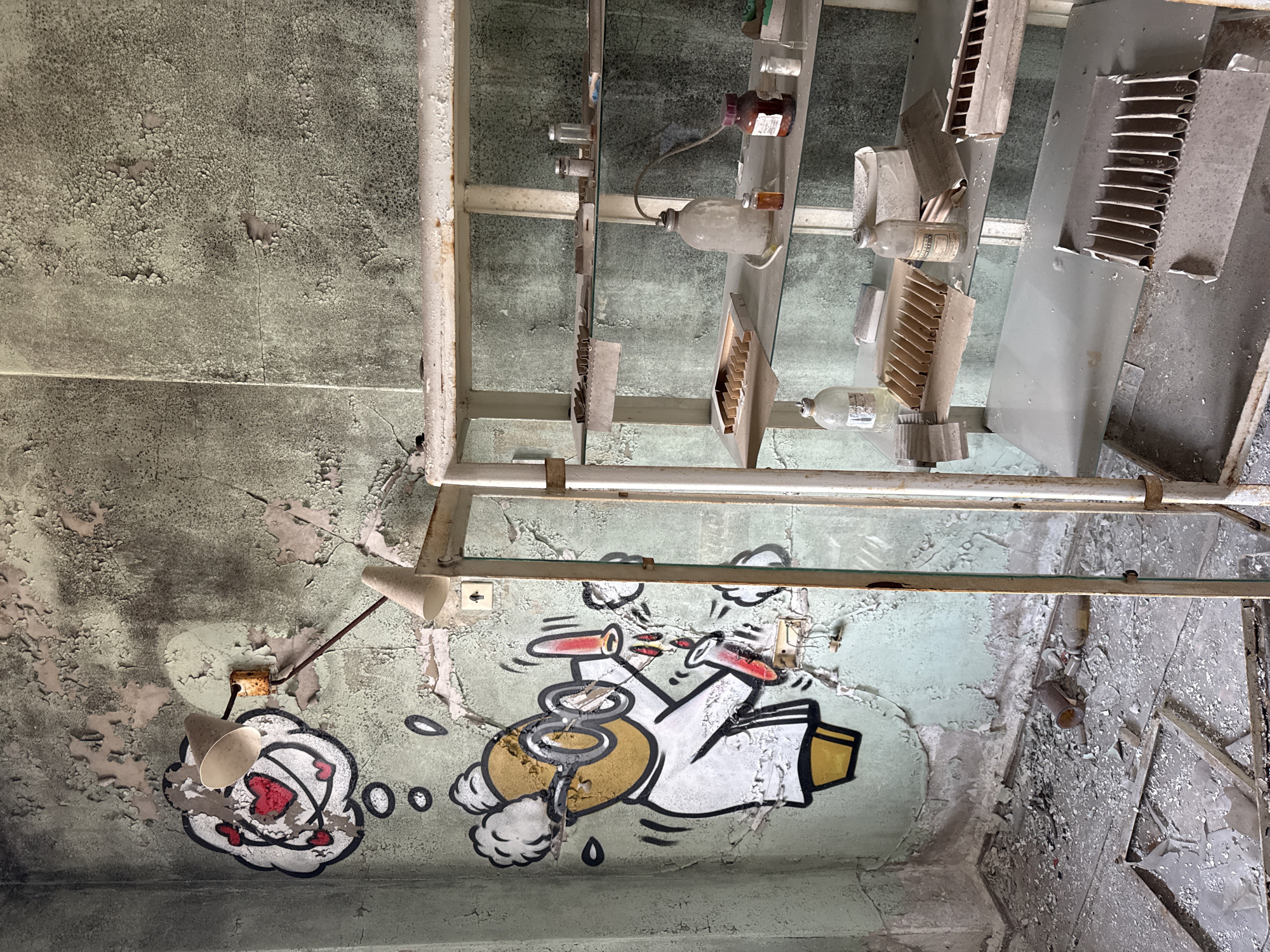 The image captures a view looking upwards in a dilapidated interior, potentially within the city of Prypiat, Ukraine. The ceiling is heavily damaged, with peeling paint and cracks. Two distinct graffiti images are visible on the ceiling. One depicts a cloud-like shape with what appear to be hearts within, resembling an atom. The other graffiti image shows a stylized depiction of laboratory equipment. Above the ceiling, a framework of metal supports is present, interspersed with various objects including bottles, vials, and ventilation components, suggesting the space may have been a laboratory or storage area within a scientific facility. The overall environment conveys a state of abandonment and decay.