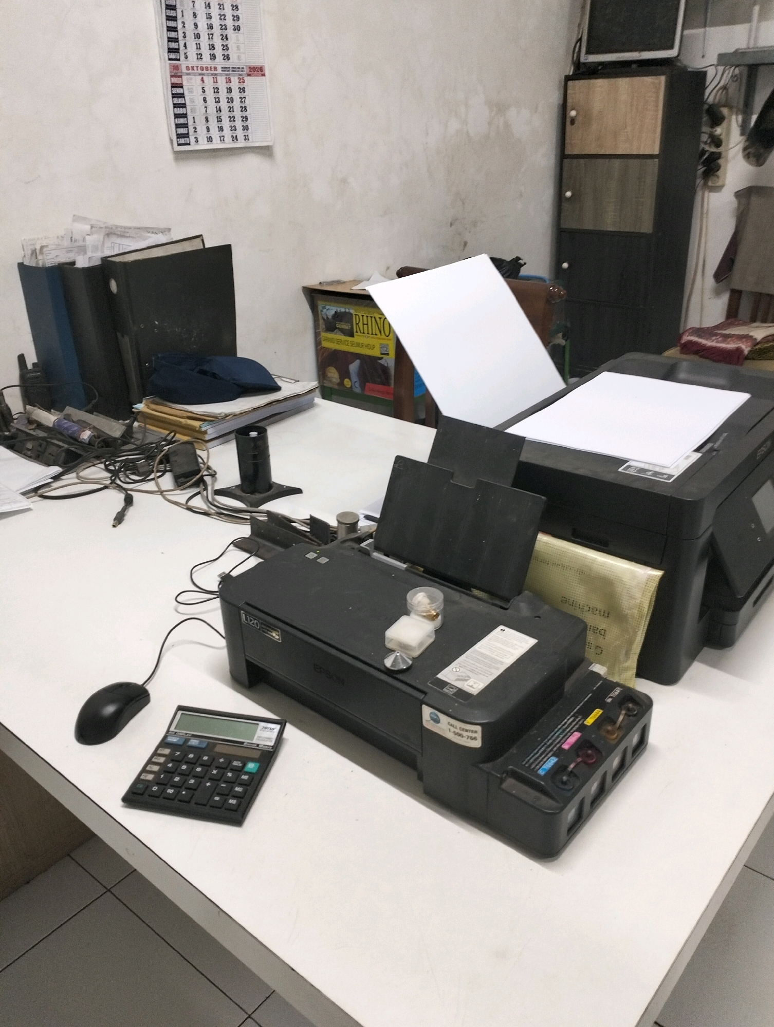The image shows a cluttered desk in what appears to be an office or home office environment. A white desk is the primary surface, with various office supplies and equipment. In the foreground, a black printer with an open lid sits on the desk, with a mouse and a black calculator to its left. Further back, a stack of folders and papers are visible. A calendar with dates for October is mounted on a textured, light-colored wall in the background. To the right of the printer, a tall, dark cabinet with three sections is visible, topped by a small electronic device. The overall impression is one of a working space with a significant amount of administrative materials and technology.