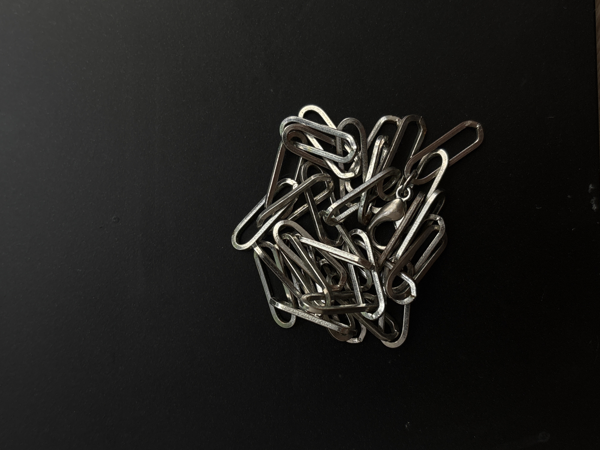 A pile of silver-colored paper clips is clustered on a black surface. The paper clips are interlinked in a tangled heap, with some lying flat and others standing on their edges. The lighting creates strong shadows, highlighting the metallic sheen of the clips and the texture of the surface. There are no people visible, and the scene appears to be indoors. The dark background and the reflective nature of the paper clips suggest a simple studio or desk setting, possibly for a product photograph or a still life study. No text is visible in the image.