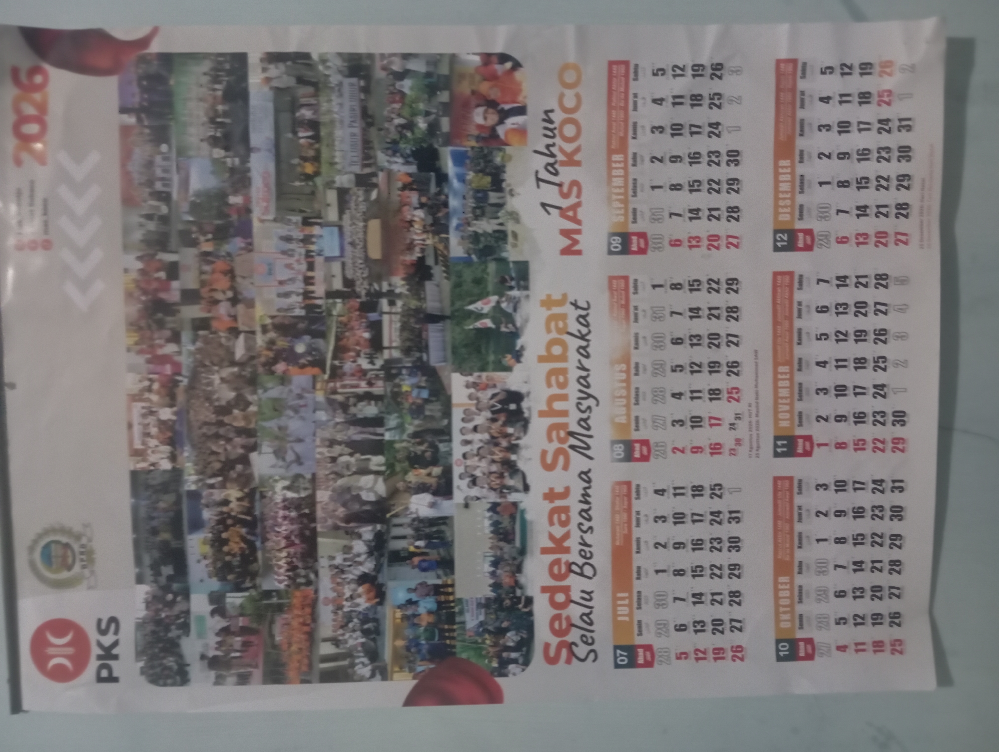 The image is a calendar for the year 2026, featuring a collage of photographs at the top. The calendar is predominantly white with text and dates in various colors, including red, black, and orange. The bottom section of the calendar is divided into monthly grids from July to December. Each month displays the days of the week (Ahad, Senin, Selasa, Rabu, Kamis, Jumat, Sabtu) and the corresponding dates. The calendar has the phrase "Sedekat Sahabat Selalu Bersama Masyarakat" printed above the monthly grids. To the right of this phrase, it says "1 Tahun MAS KOCO". The PKS logo and another emblem are visible at the top left of the calendar. The background is a light, neutral color, likely a wall. The image is taken from a slightly downward angle. The overall impression is that of a political or organizational calendar. No specific details about Kutasari, Indonesia, can be inferred from the image itself.