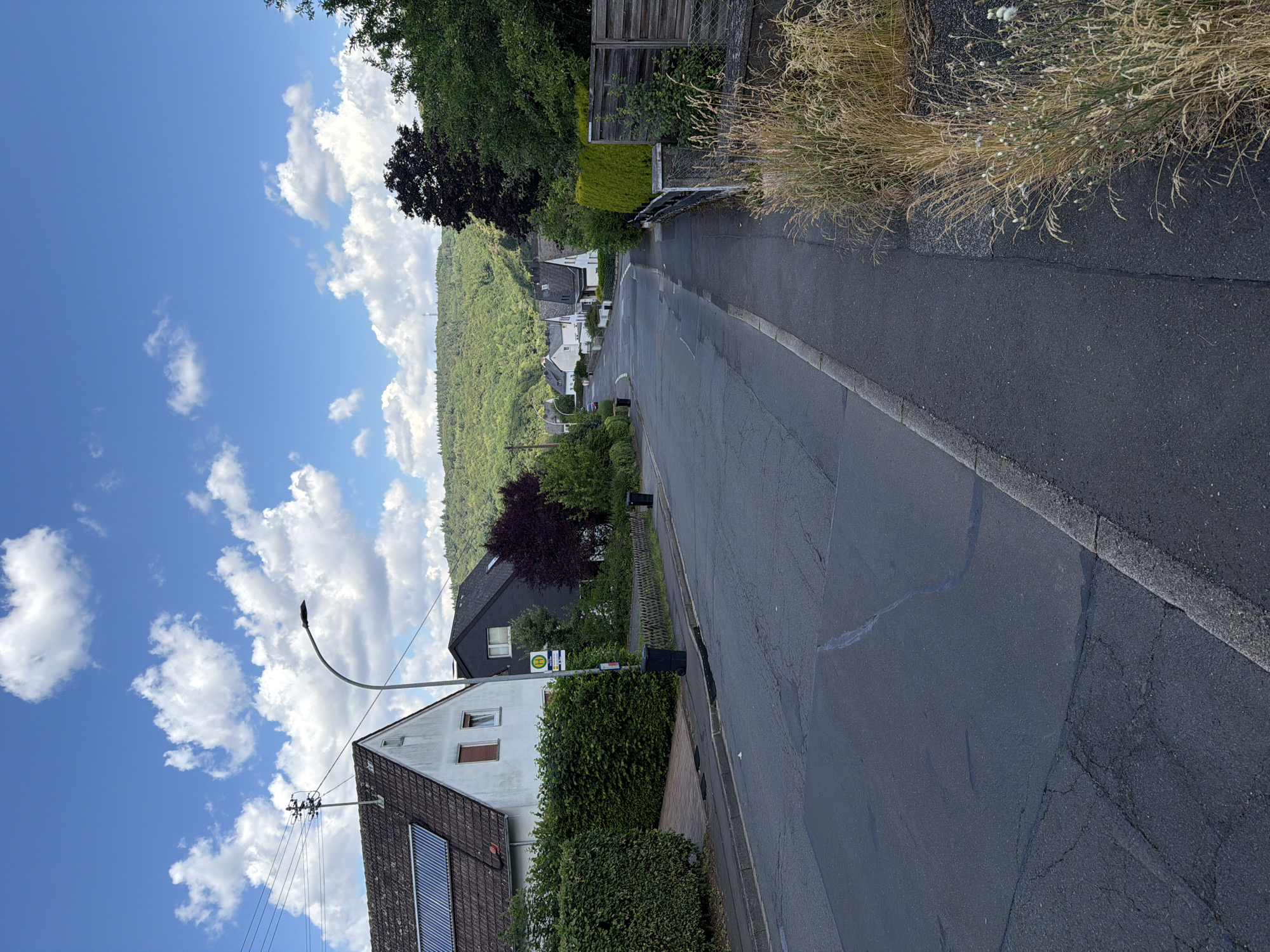 The image depicts a street in Idar-Oberstein, Germany, on a clear day with a blue sky filled with fluffy white clouds. The street is paved and appears to be in a residential area, with houses lining both sides. On the left, a white house with solar panels on its roof is visible, adjacent to a neatly trimmed green hedge. Further down the street, a bus stop sign with a green 'H' symbol indicates public transportation availability. A dark gray house with a sloped roof stands next to the white house. The right side of the street features a sidewalk and a wooden fence separating the properties from the road. Tall, dry grasses and some flowering plants grow along the edge of the sidewalk. In the background, a lush green hillside covered in trees rises up, with a few more houses scattered along its lower slopes. The overall impression is of a quiet, suburban street scene.