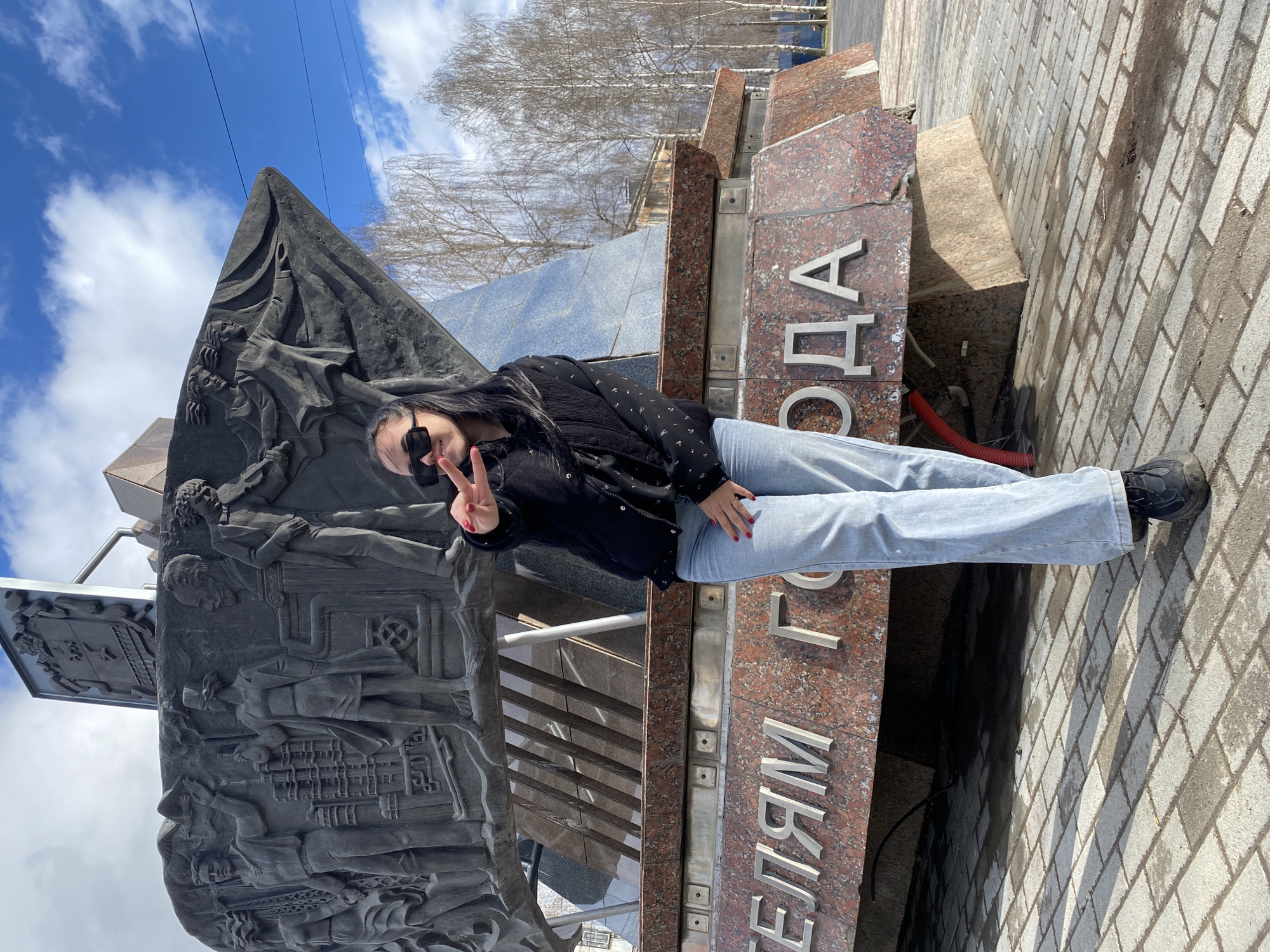 A young woman is captured posing for a photo next to a prominent monument in Chaikovsky, Russia, on a bright, sunny day with a blue sky dotted with white clouds. She stands casually, facing the camera, wearing dark sunglasses, a black patterned jacket, light blue jeans, and black shoes. Her left hand is raised, forming a peace sign, while her right hand rests near her face, holding the sunglasses.
The monument beside her is multifaceted. To her left, there's a tall, dark-colored structure, possibly made of bronze or dark stone, featuring intricate bas-reliefs depicting various figures and architectural/industrial scenes, suggesting a commemorative or historical theme. To her right, a section of the monument is made of polished red and black speckled granite, upon which large, silver metal letters are affixed. Visible portions of the text read "ЕЛЯМ" and "ГОДА," which likely form part of the phrase "СТРОИТЕЛЯМ ГОРОДА" (To the Builders of the City), a common dedication for monuments in Russian cities.
In the background, bare trees indicate an early spring or late autumn season. The ground is paved with light-colored brick-like tiles, creating a well-maintained urban setting. The overall mood of the image is cheerful and relaxed, reflecting the woman's pose and the pleasant weather.