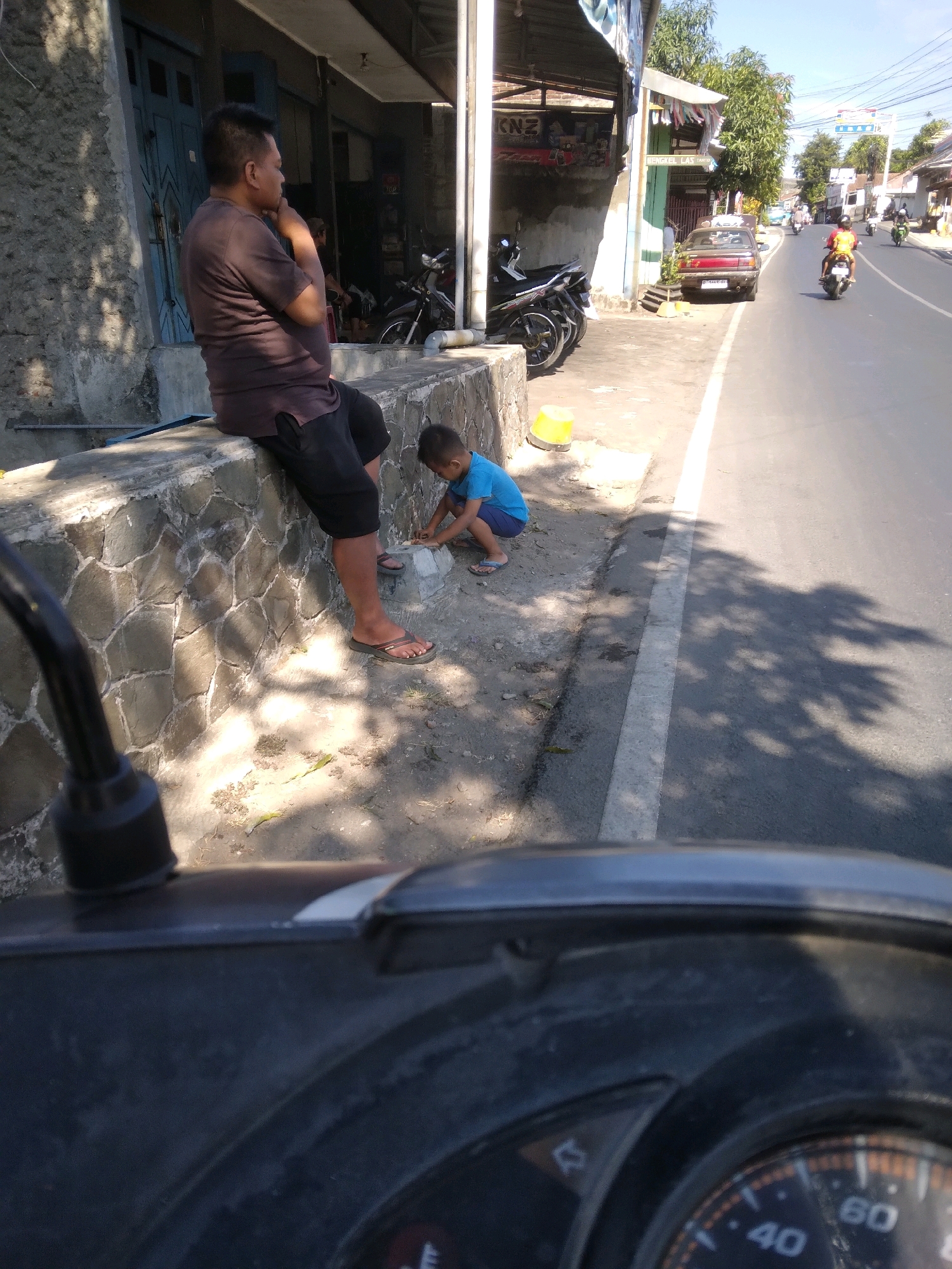 In Kedungmalang, Indonesia, a man in a dark t-shirt and shorts sits on a low stone wall. Next to him, a young boy in a blue t-shirt and shorts crouches on the ground, playing with something near the wall. A yellow bucket is visible behind the boy. The scene takes place on a street with a paved road and a sidewalk. Several motorcycles are parked in front of a building to the left, and a car is parked on the side of the road further down. The sun casts strong shadows across the ground and road. The foreground of the image is filled with the dashboard of a scooter.