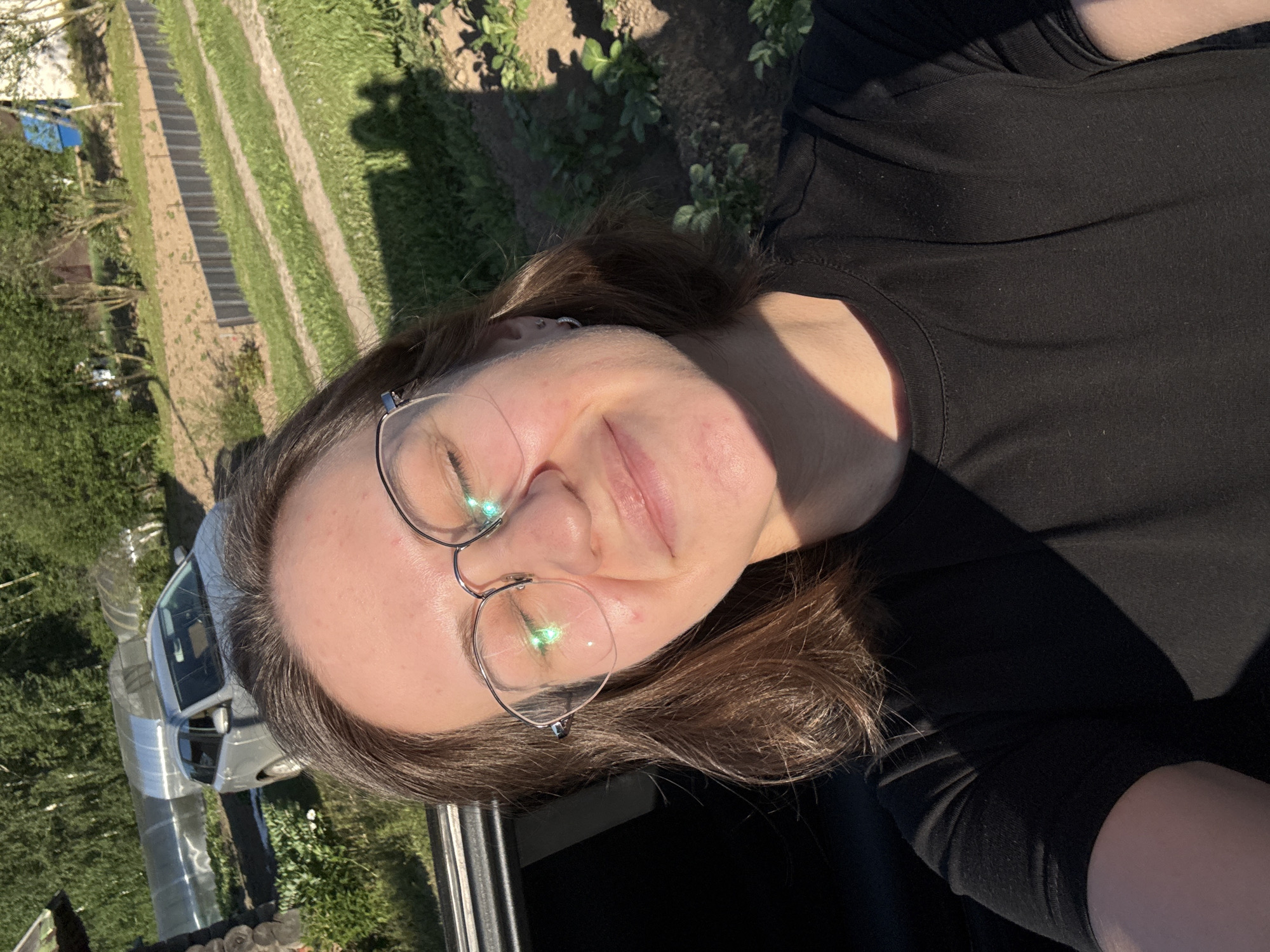 A young adult female is depicted in a close-up outdoor selfie, situated in Lenino, Russia, under strong, direct sunlight. She has shoulder-length brown hair, a fair complexion with minor facial blemishes, and wears round, metal-rimmed glasses, which reflect green light. Her eyes are closed or squinting, and she has a subtle smile. A small silver hoop earring is visible in her right ear, along with a second, higher piercing. She is wearing a black crew-neck t-shirt, largely in shadow from the sun coming from the upper right, casting a distinct shadow across her torso.
Behind her on the left, part of a dark car interior or door frame is visible, along with a section of a rough-hewn wooden log structure. Further in the background, a silver/light grey station wagon is parked on unpaved ground, and two long, tunnel-style greenhouses with clear plastic coverings are visible, one appearing somewhat collapsed. The ground features strips of green grass, tilled earth, and dirt tracks. A low, dark grey corrugated metal fence runs horizontally across the mid-ground. To the right, shadows of at least two individuals are cast on the ground among rows of small green leafy plants, possibly a garden crop. Dense green trees and bushes form a natural backdrop in the distance, with a small structure featuring a blue wall/roof section visible on the far right.