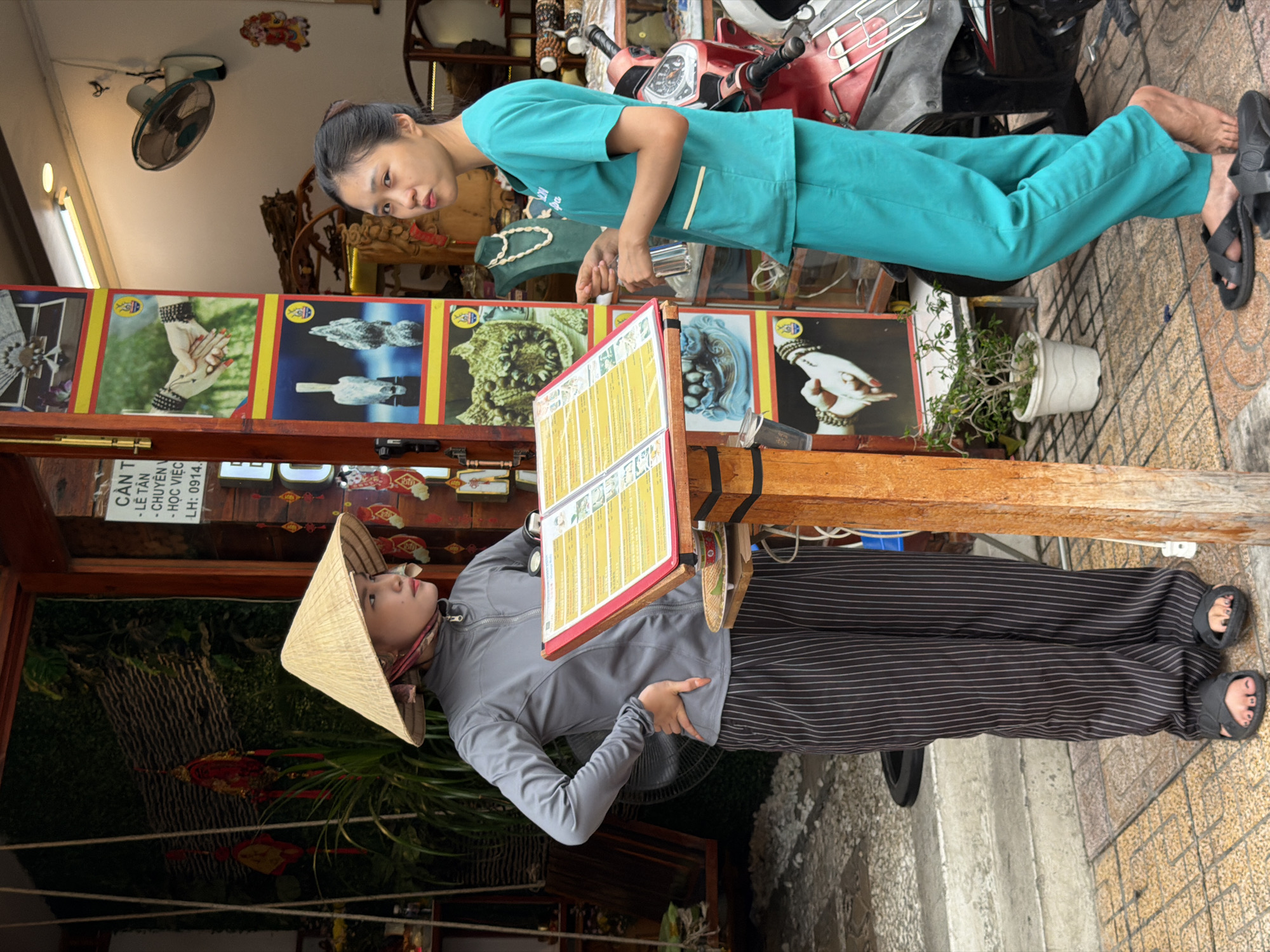 Two women are standing outside of a shop in Nha Trang Ward, Vietnam. One woman is wearing a conical Asian hat, a gray jacket, and striped pants. She is standing behind a menu and leaning against a wooden stand. The other woman is wearing a teal uniform and sandals. A scooter is parked behind her. The shop has framed posters above the menu. A sign with Vietnamese writing hangs above the shop entrance. A ceiling fan is visible. The ground is tiled.