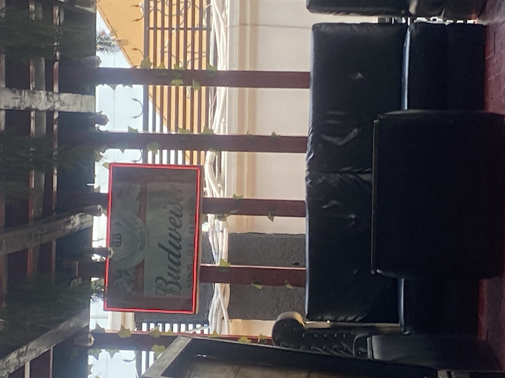 The image shows an outdoor seating area under a wooden pergola. A red-lit sign with the Budweiser logo is prominently displayed, hanging from the pergola. Below the sign, a dark leather sofa is visible, with a dark rectangular table in front of it. The background reveals horizontal slatted fencing and a building with an orange roof. The scene is set in Howayi, Nigeria. There are no people visible in the image.
