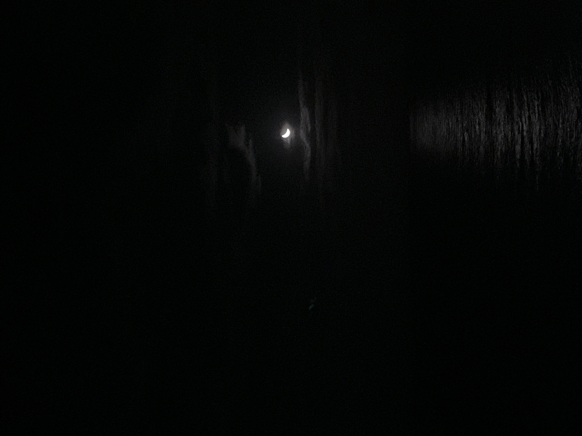 The image depicts a dark, nighttime scene. A crescent moon is visible amidst clouds, providing the primary source of illumination. To the right, a darker area with a linear texture suggests trees or foliage. The location is Unknown, Unknown.
