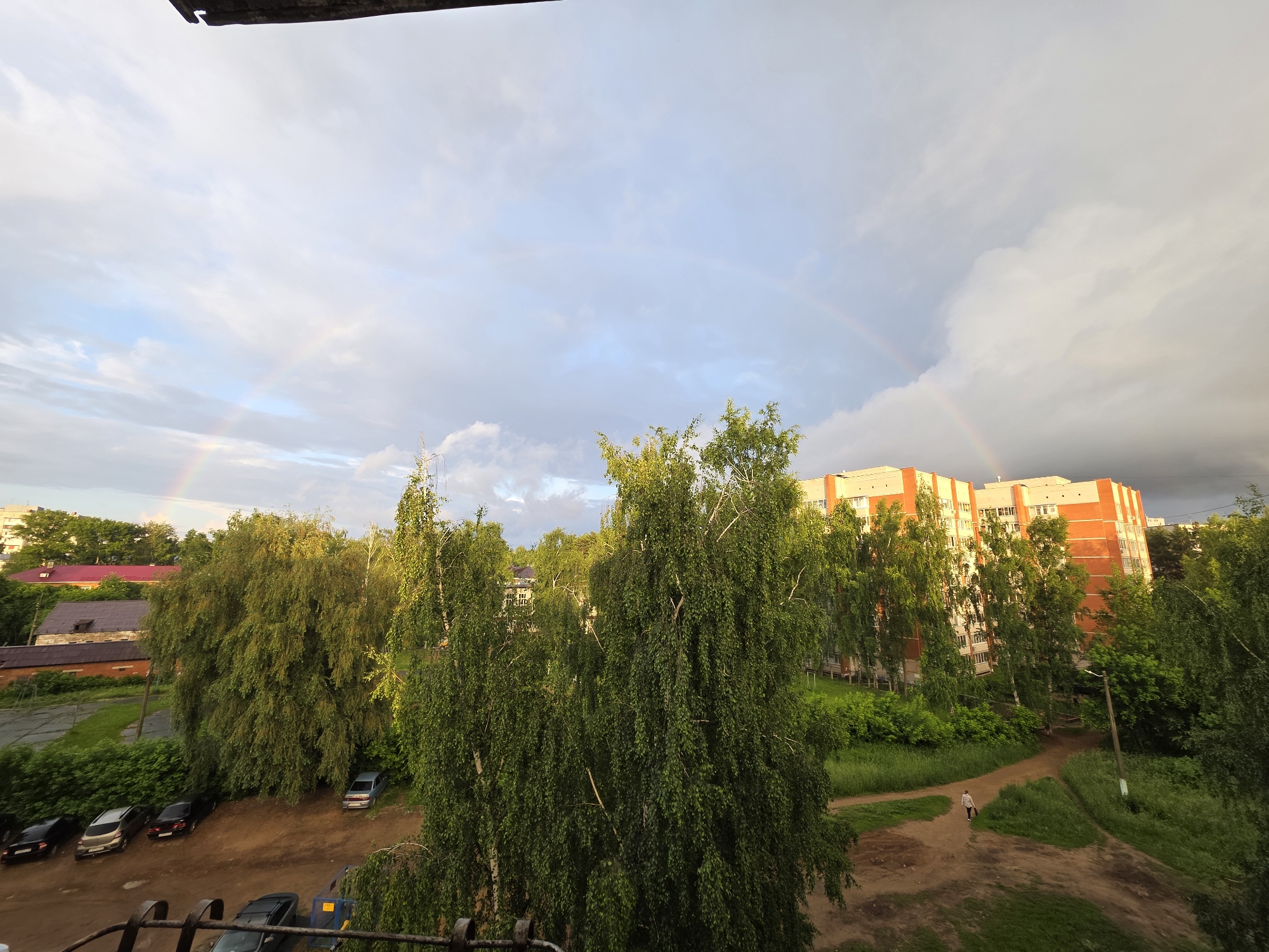 The image depicts a rainbow arcing across a cloudy sky over a residential area in Chaikovsky, Russia. In the foreground, a dark metal railing is visible, suggesting the vantage point is from a balcony or window. Beyond the railing, lush green trees dominate the middle ground, with a large, dense birch tree in the center and other trees surrounding it. To the left, several parked cars are visible on a dirt and gravel area. Further back, several multi-story apartment buildings with orange and white facades are situated amidst more greenery. A single person is walking on a dirt path in front of the buildings. The sky is a mix of light blue and heavy gray clouds, with the rainbow clearly visible, indicating recent rainfall or ongoing showers. The overall scene captures a tranquil moment in a Russian urban landscape following a weather event.