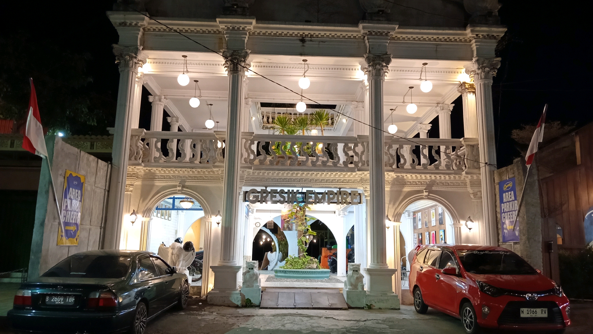 At night, in front of a white building with ornate columns and a balcony, a red car is parked on the right, and a dark green car is parked on the left. The building's name, "GRESIK EMPIRE," is visible in illuminated letters above the entrance. Two flags, one red and white and the other blue and yellow with text, are mounted on poles to the left and right of the building's facade. The area is lit by numerous hanging globe lights, and two small stone lion statues flank the central pathway leading to the entrance. The setting appears to be a commercial establishment in the city of Gresik, Indonesia.